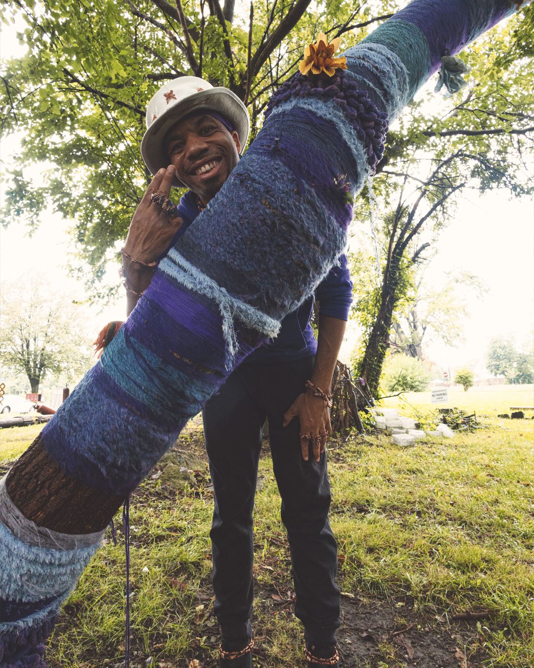 Man hugging a yarn-bombed tree wrapped in colorful knitted fabric in a park, smiling and flashing a peace sign.