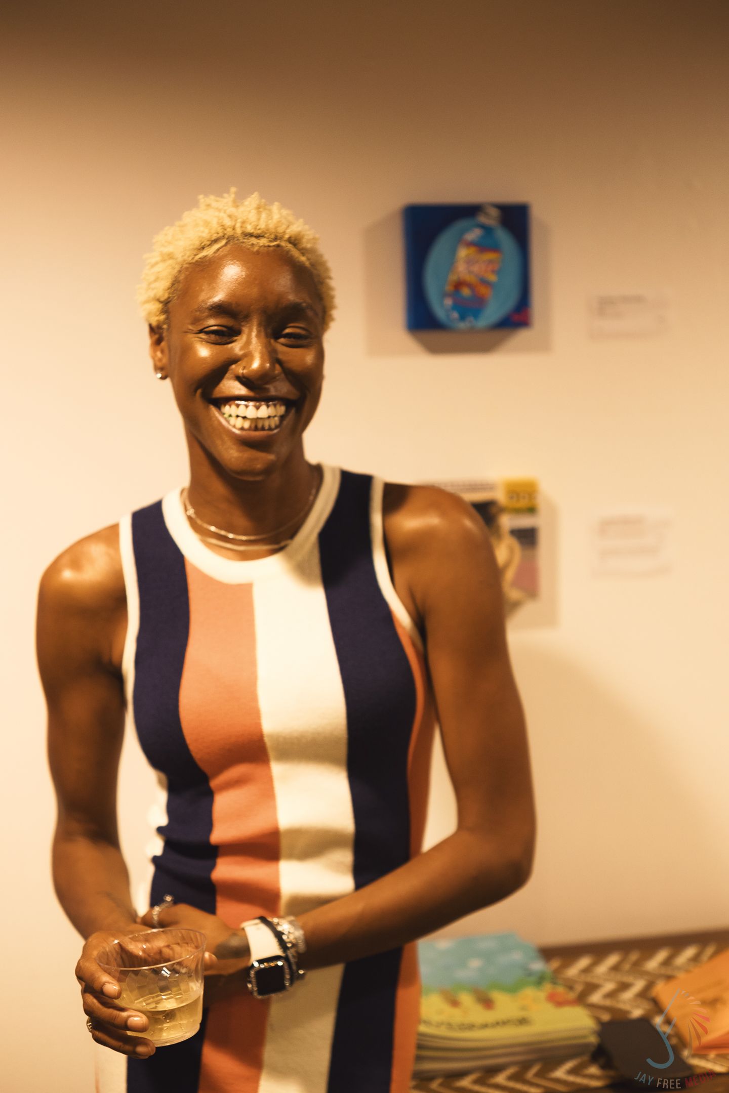 Smiling woman with blonde hair wearing striped dress holds drink at art gallery event.