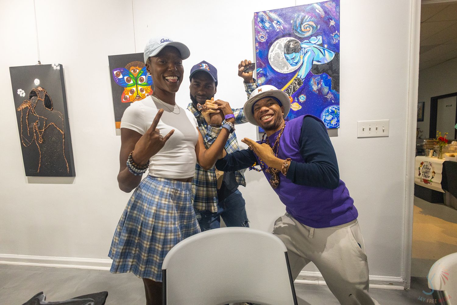 Three friends pose and smile together at an art gallery with colorful paintings displayed on the walls behind them.