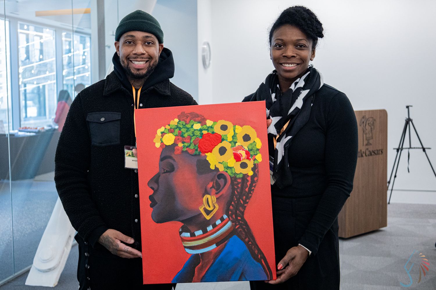 Two people proudly display a vibrant painting of an African woman with floral headpiece at an art event.