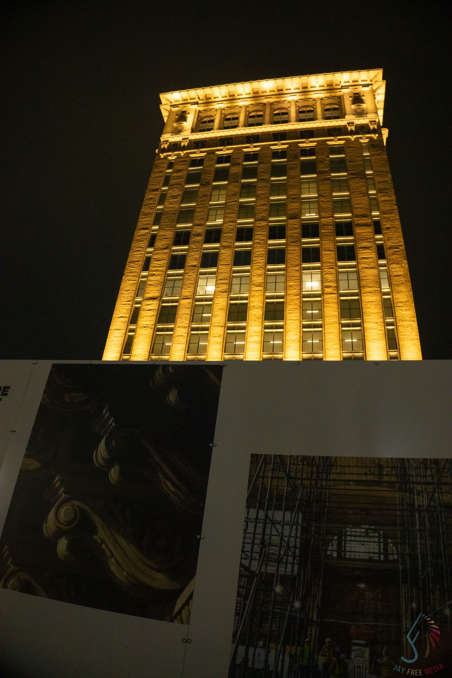 A tall historic building illuminated in golden light at night with construction photos displayed in foreground.