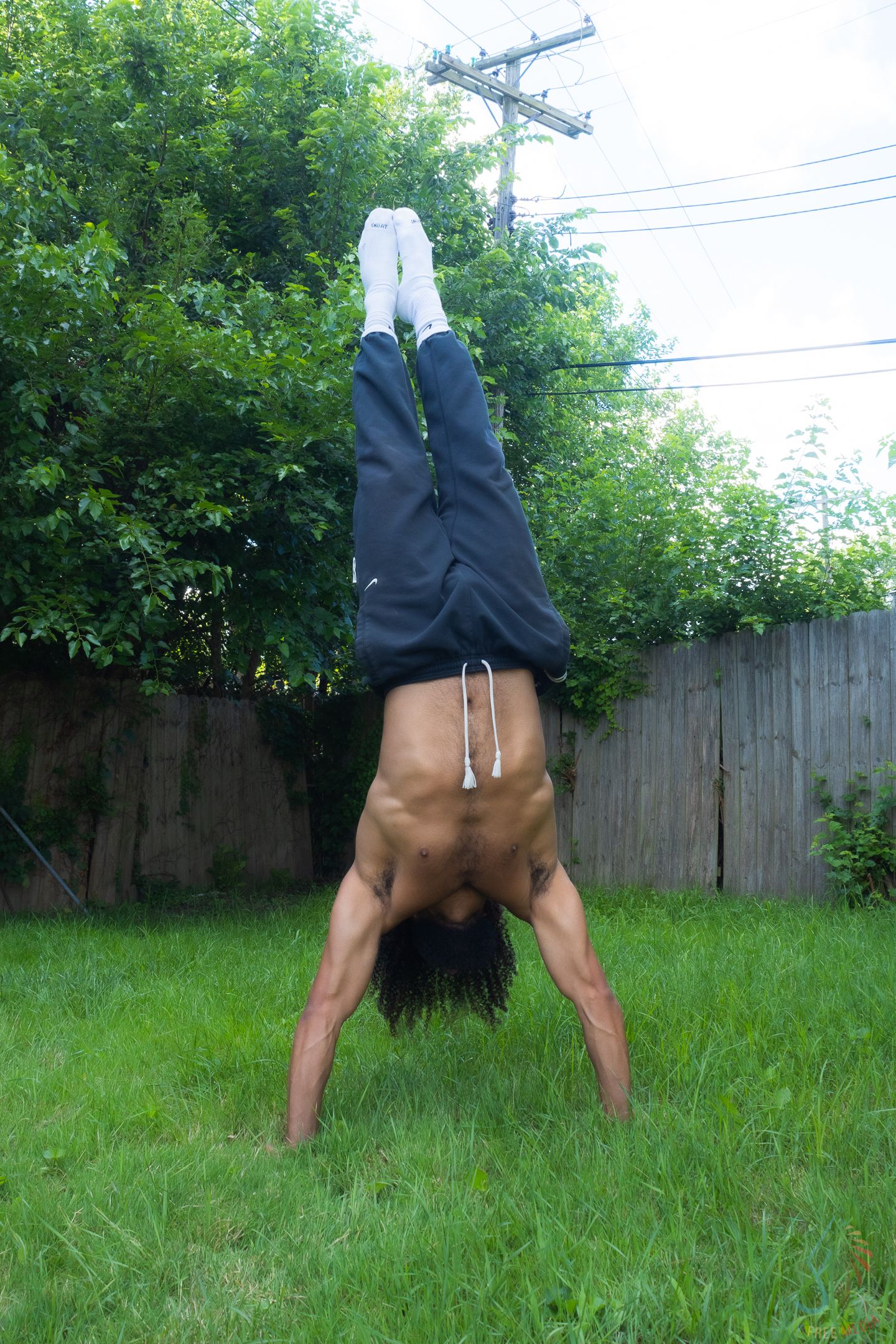 Shirtless man performing a handstand on green grass in a backyard, wearing dark sweatpants and white socks.