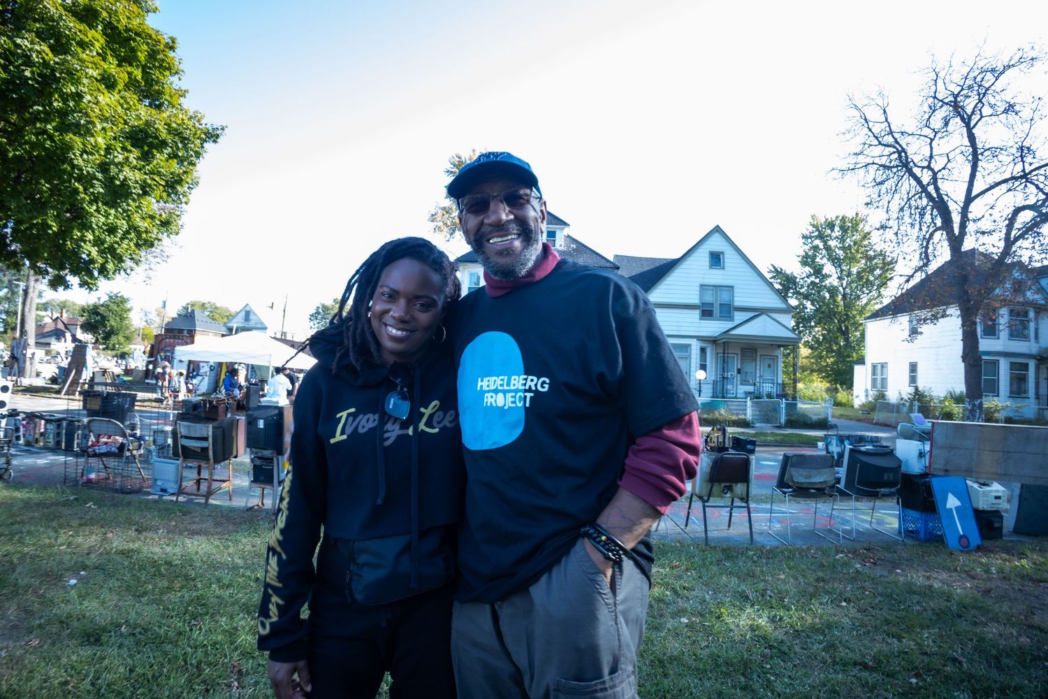 Two people smiling and posing together outdoors at a community event, wearing matching Heidelberg Project sweatshirts.