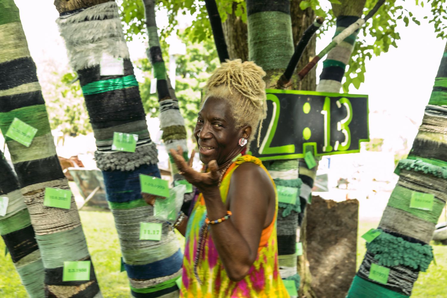 A woman with dreadlocks smiles among yarn-bombed trees decorated with green knitted wraps and tags outdoors.