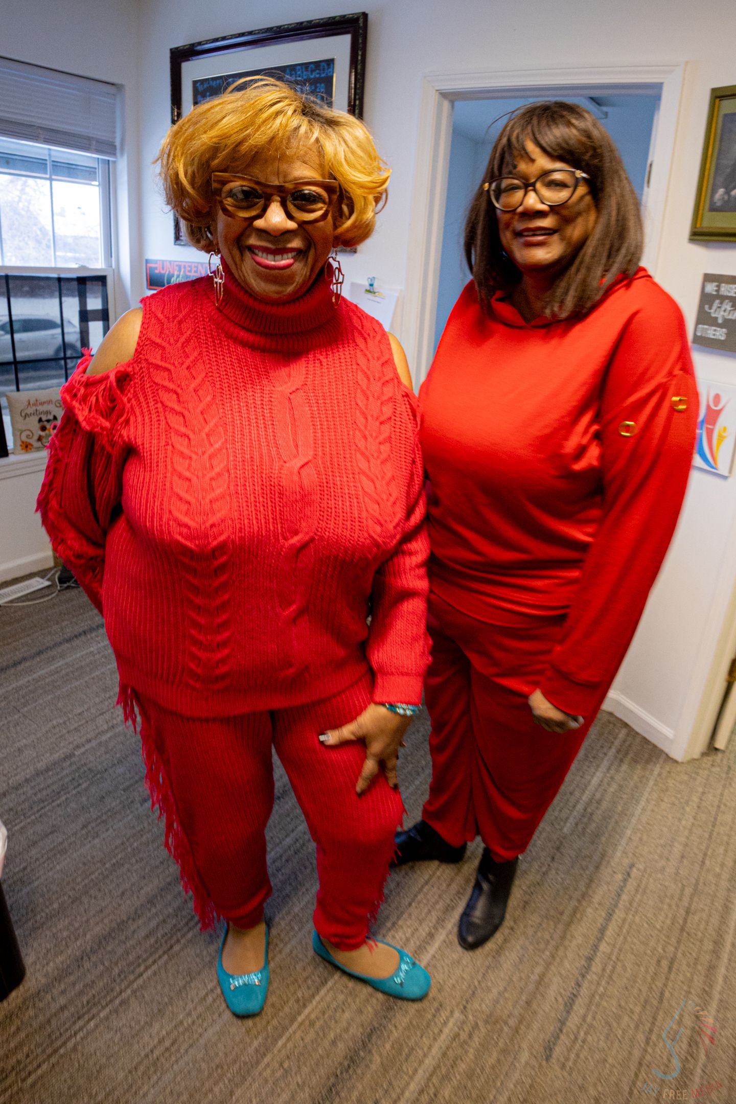 Two women wearing matching red outfits pose together smiling indoors, both accessorized with teal blue flats.