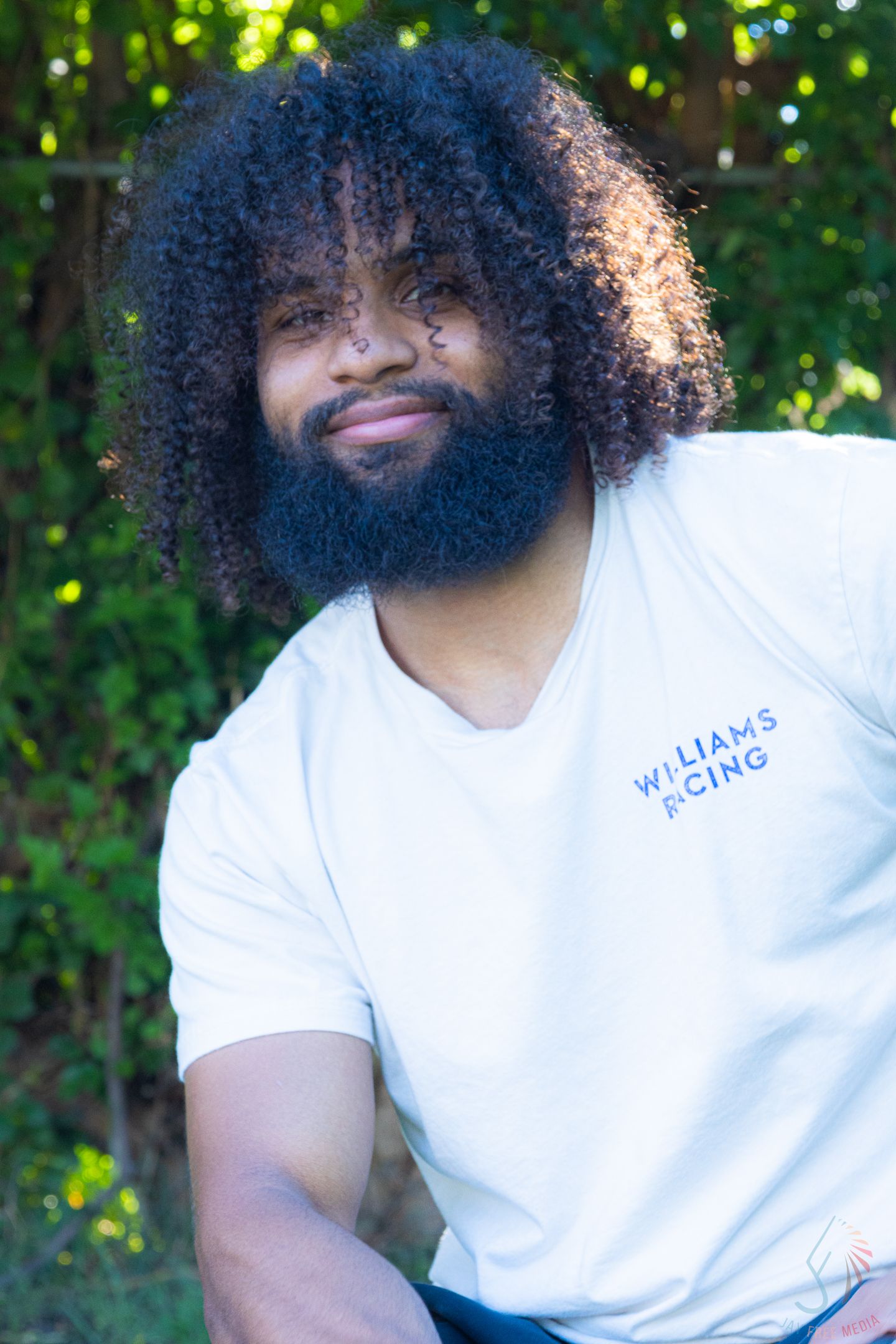 Smiling Black man with large curly afro and beard wearing a white Williams Racing t-shirt outdoors.
