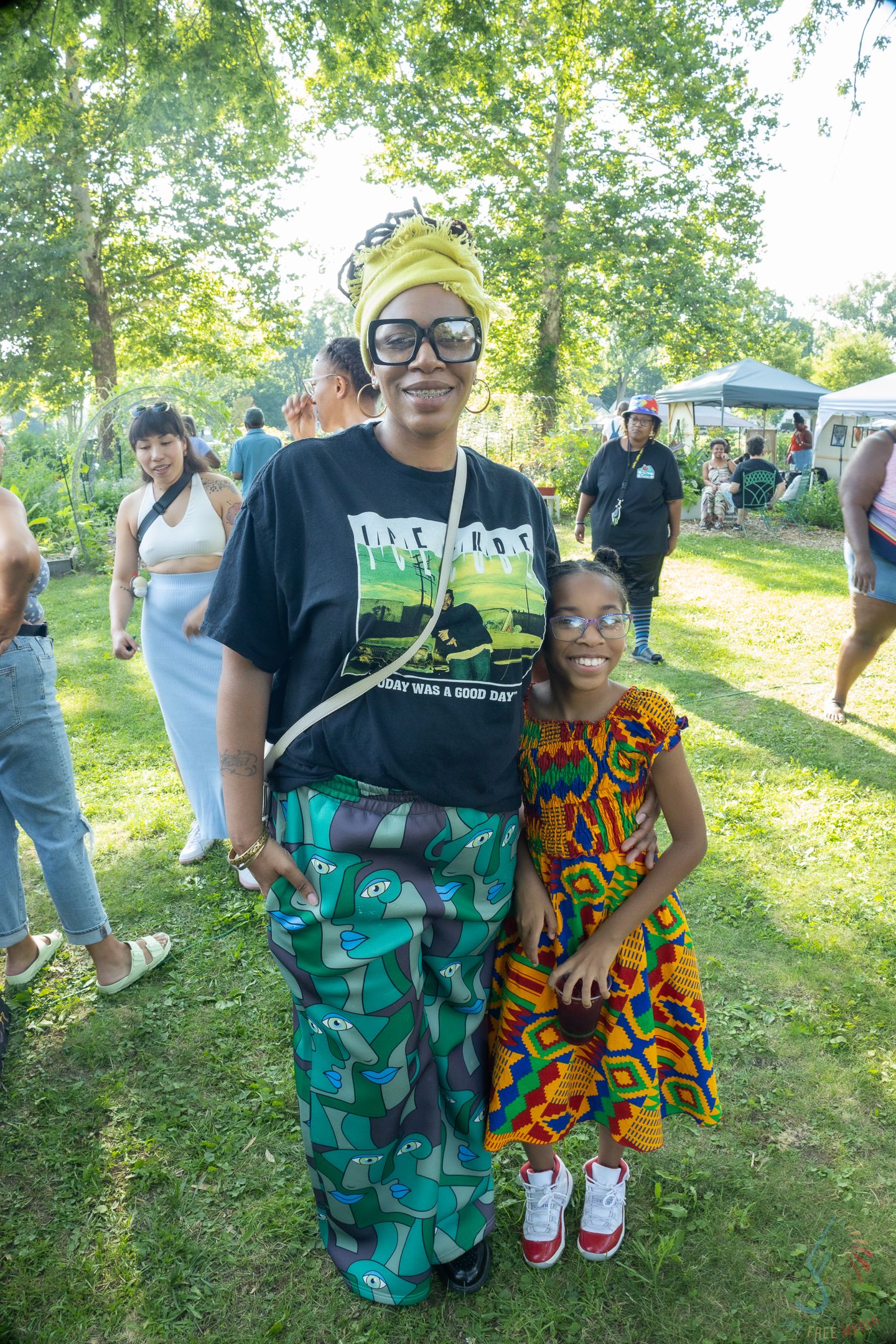Woman and young girl in colorful African print outfits smiling at an outdoor cultural festival.