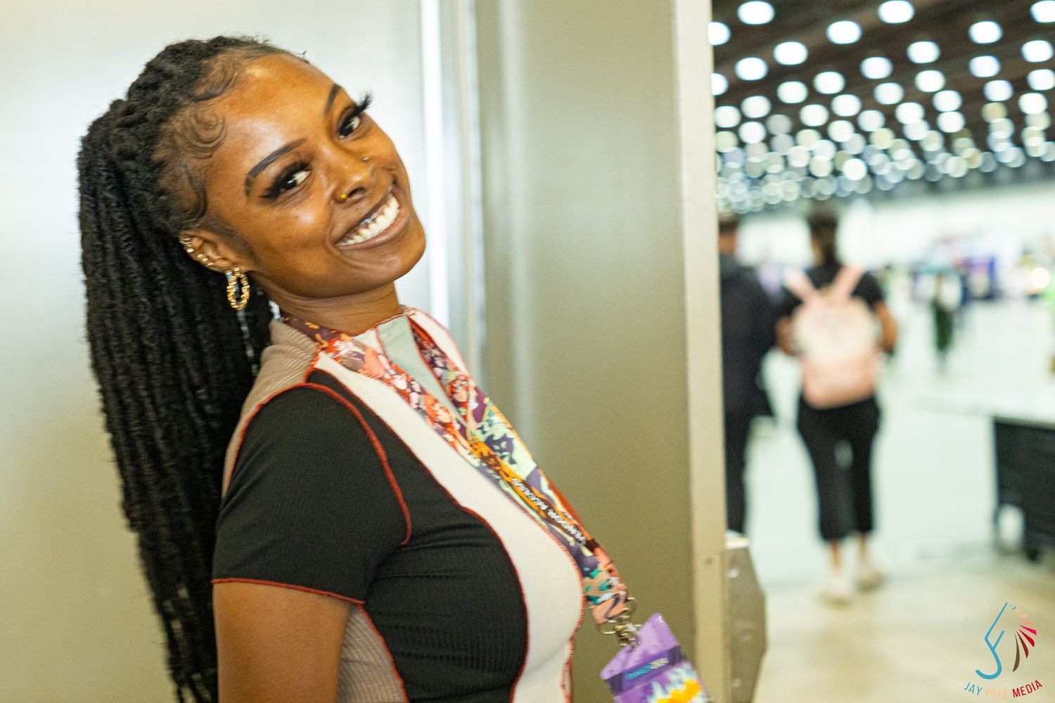 Smiling woman with long braids wearing a colorful lanyard poses at an indoor convention event.