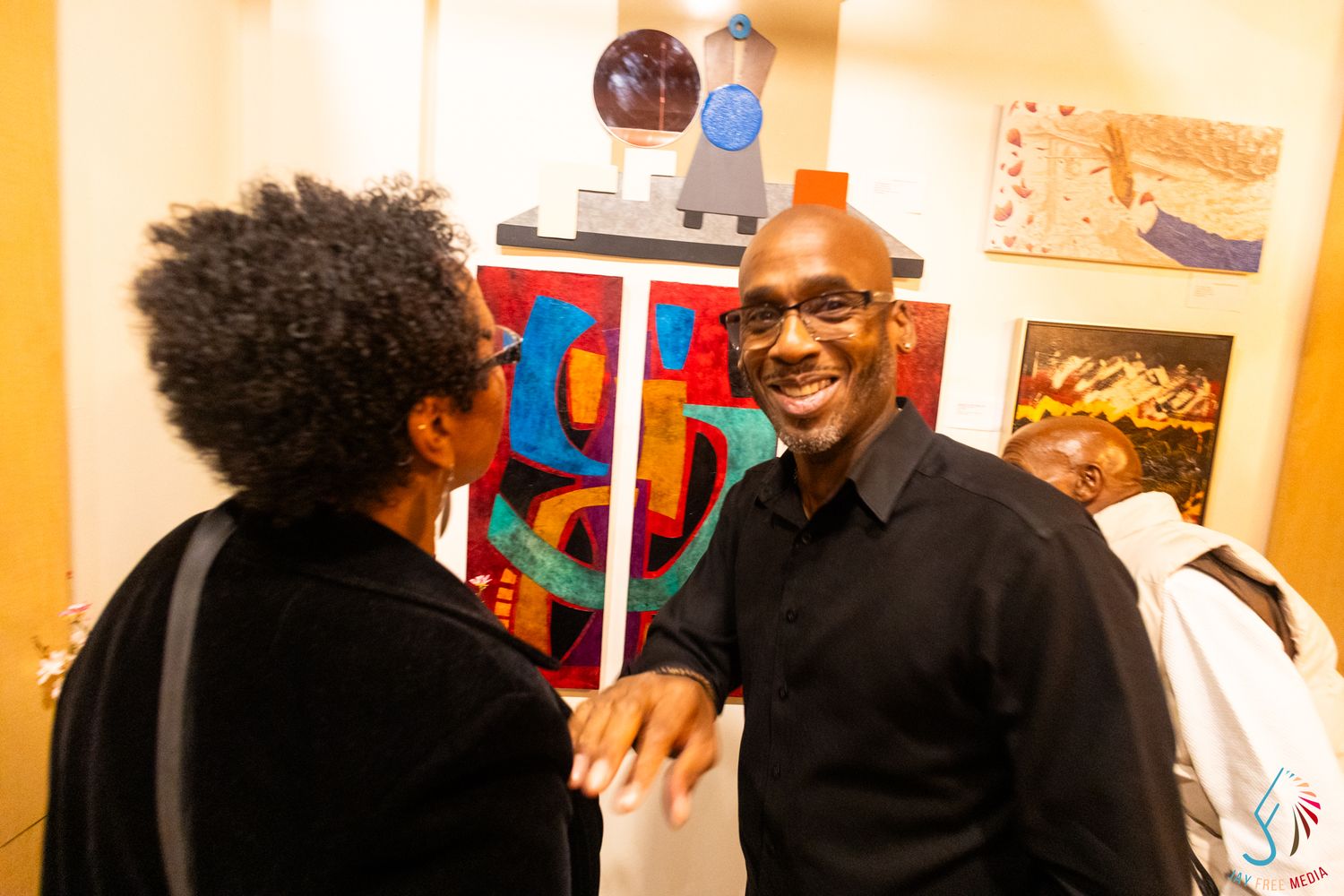 Two people smiling and shaking hands at an art gallery event with colorful paintings displayed on the wall behind them.