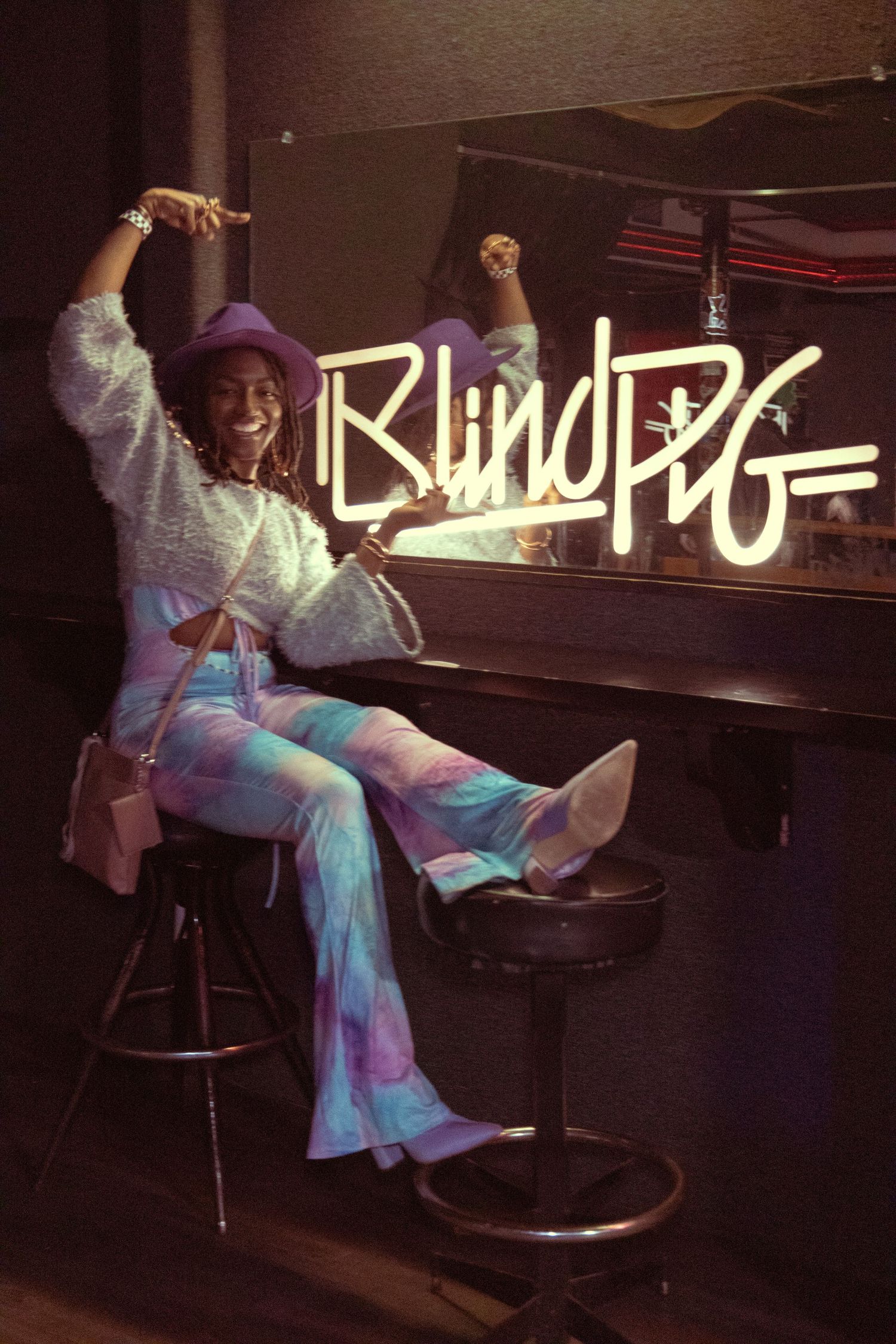 A stylish woman poses on a bar stool in front of a neon Blind Pig sign, wearing tie-dye pants and a purple hat.
