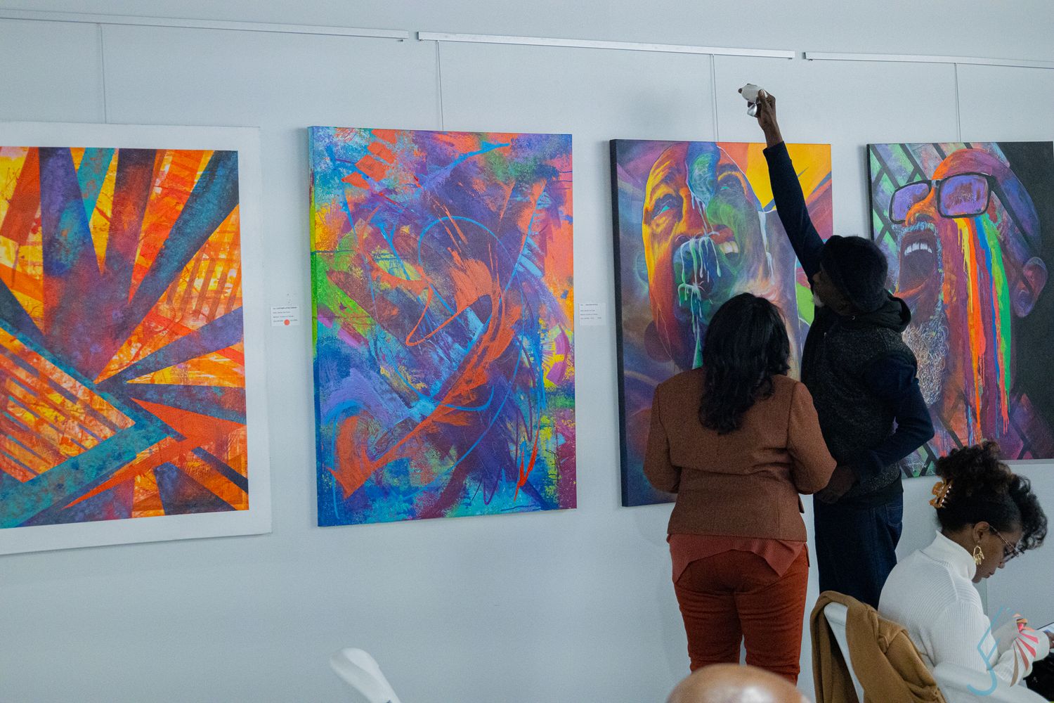 People viewing colorful abstract paintings displayed on a white gallery wall during an art exhibition.