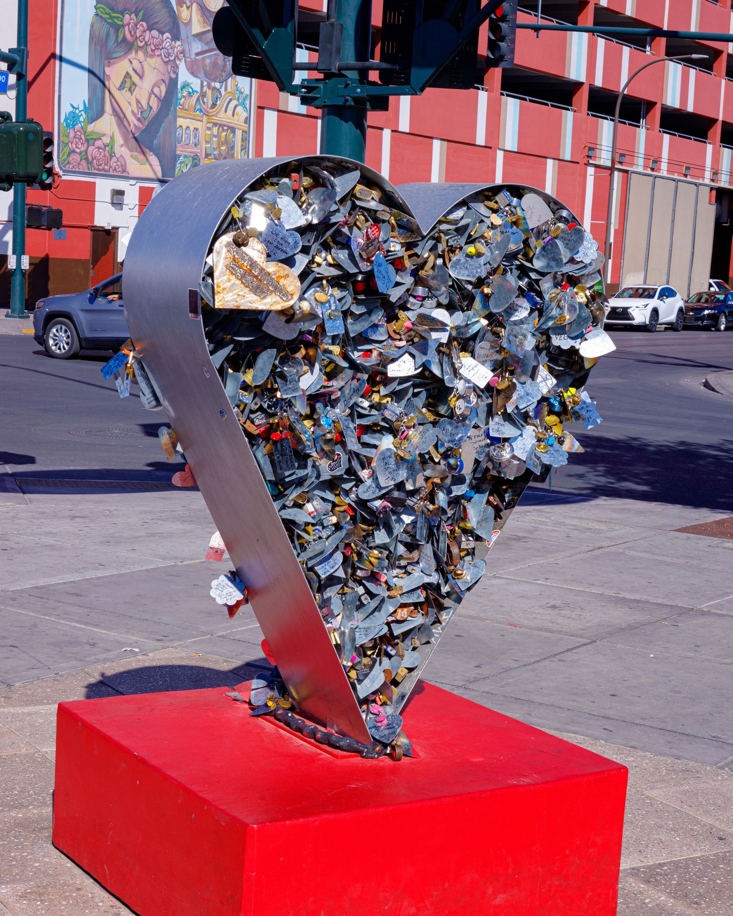 Metal heart sculpture filled with colorful locks on a red pedestal at a city intersection.