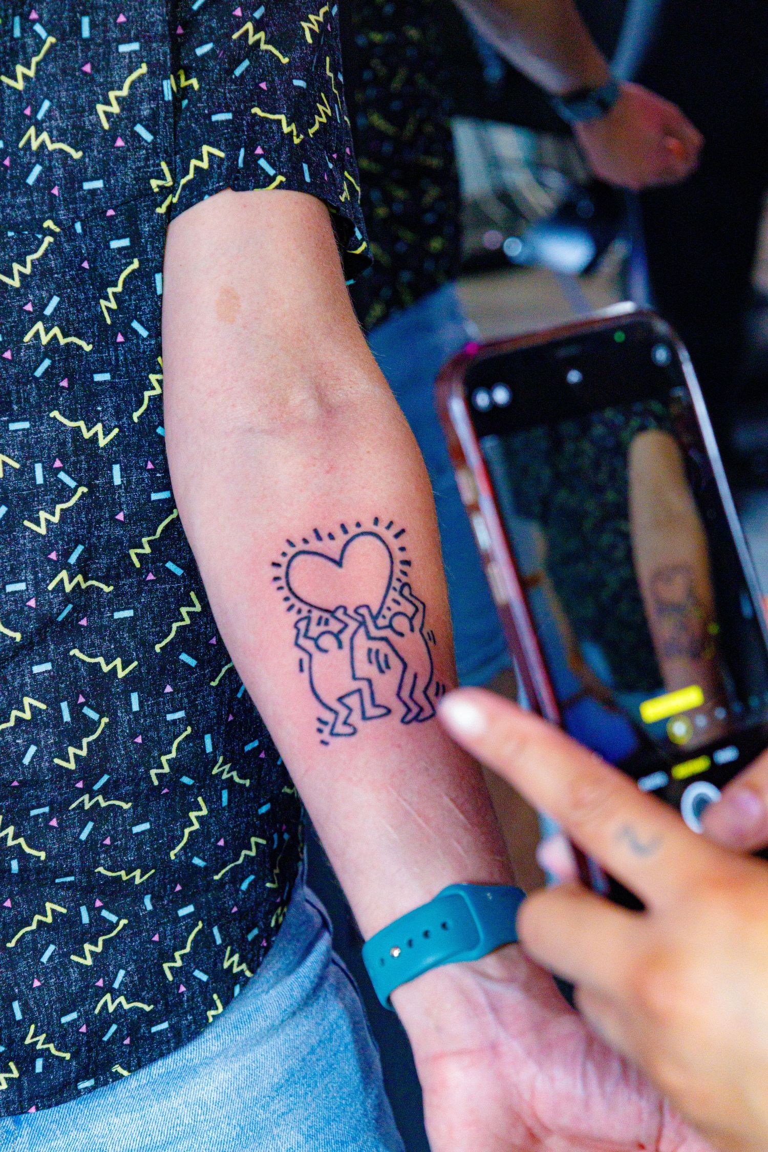 A Keith Haring inspired heart figures tattoo on a forearm being photographed with a smartphone.