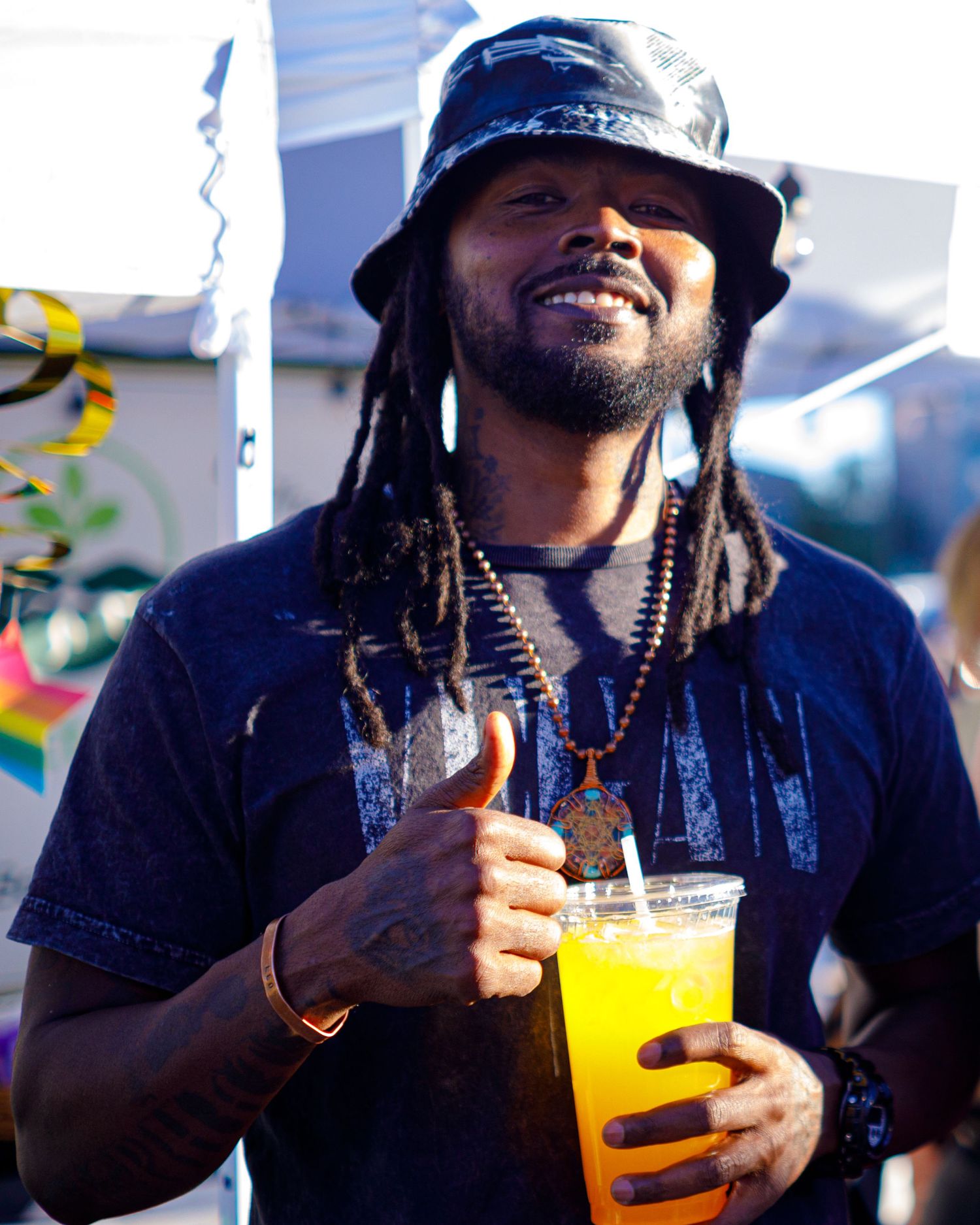 Smiling man with dreadlocks holding a orange juice drink at an outdoor festival event.