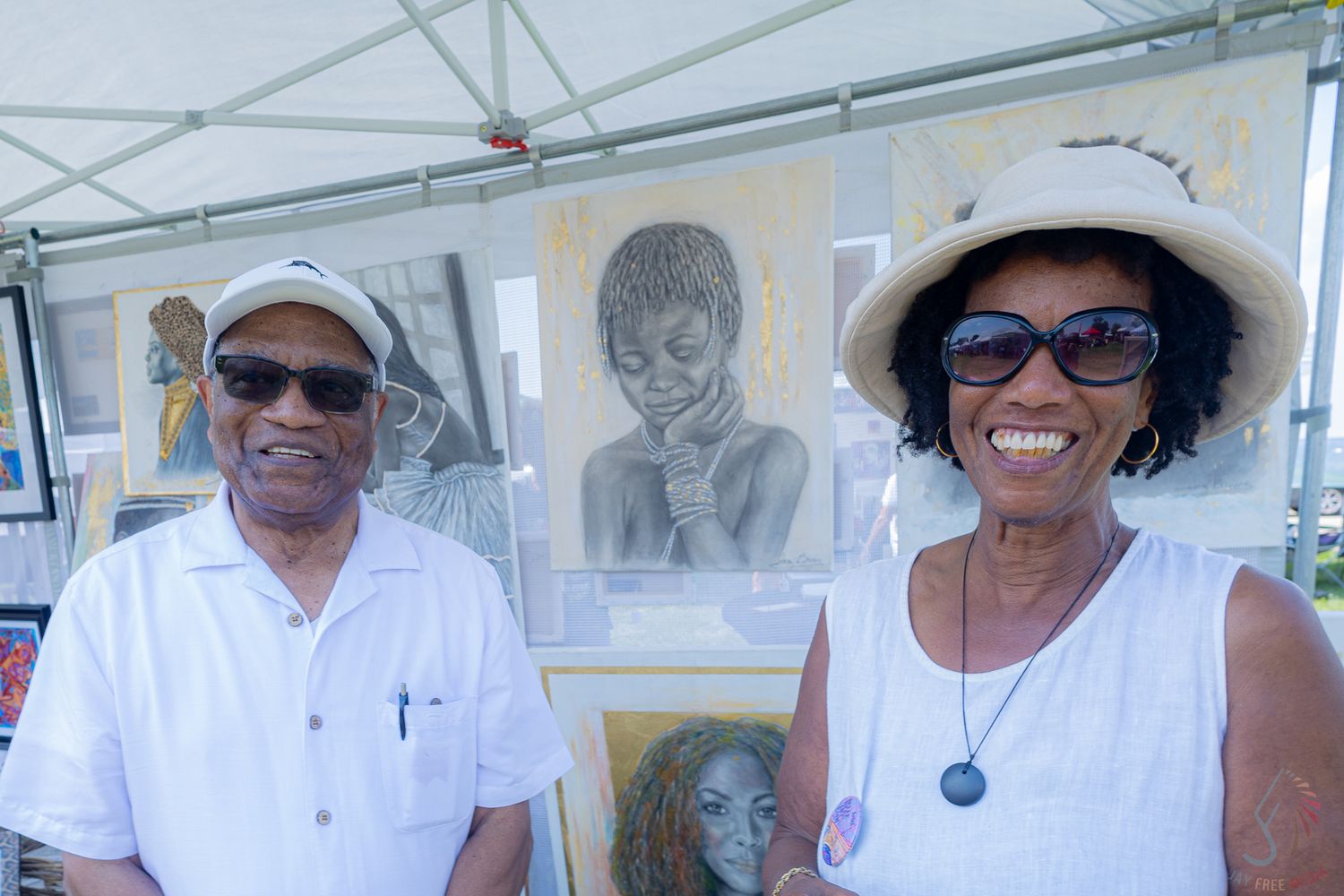 Two smiling people pose in front of African portrait artwork at an outdoor art fair.
