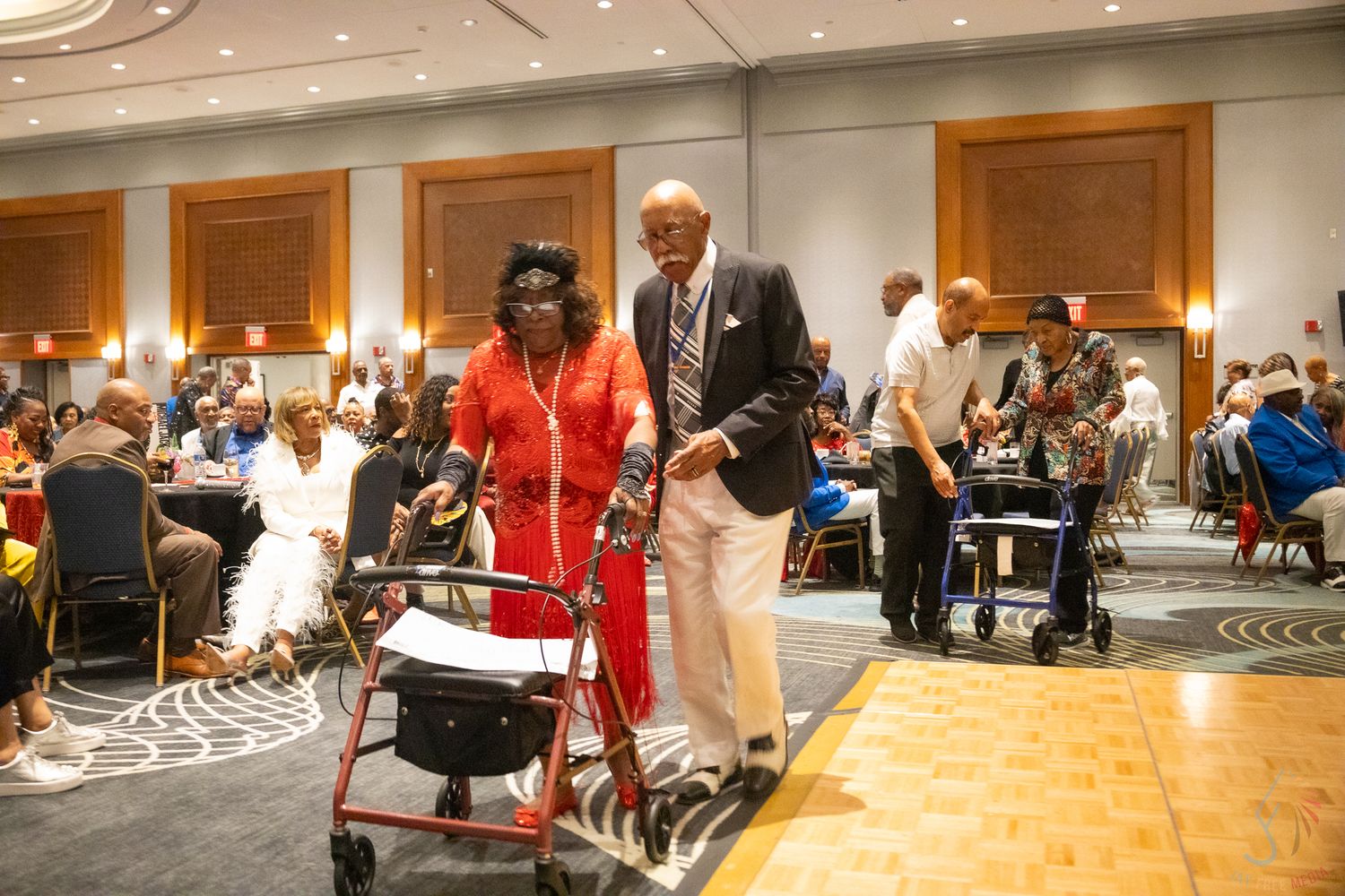 Elderly woman with walker escorted by man in white suit at a formal banquet event.