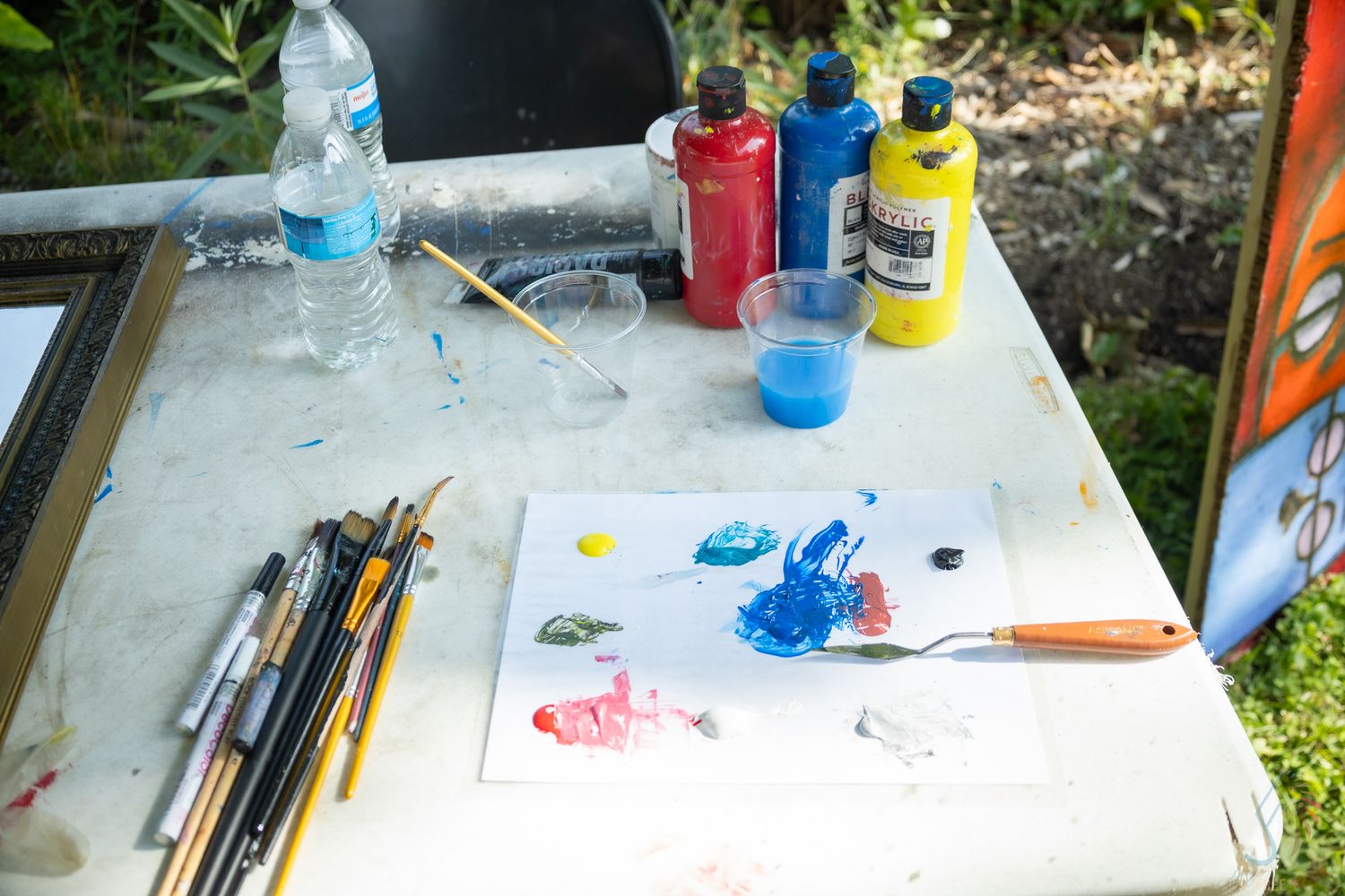 Outdoor painting setup with brushes, palette knife, red and blue acrylic paints, water bottle, and colorful paper palette.