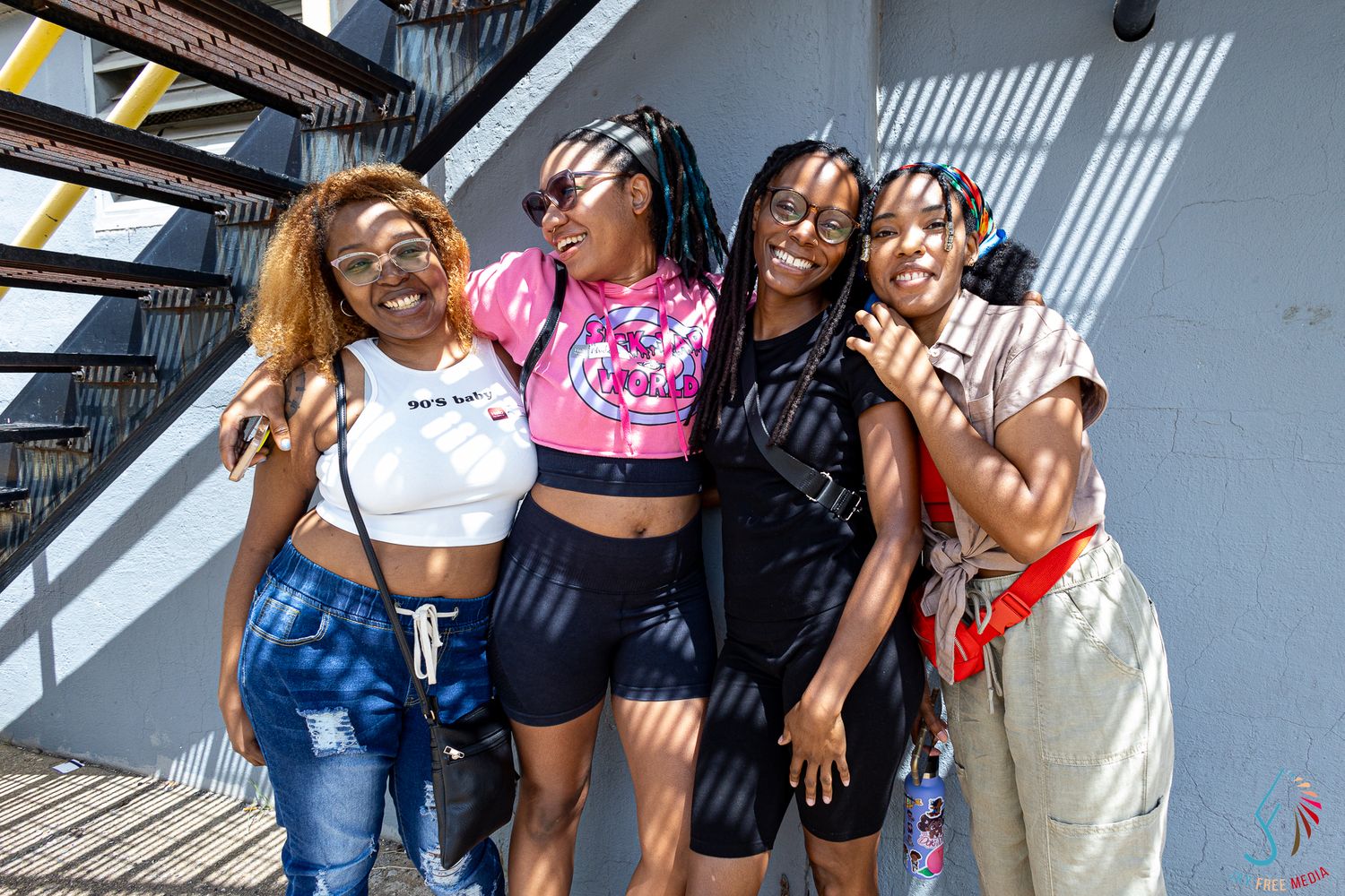 Four young women smile and pose together outdoors on a sunny day near a metal staircase.