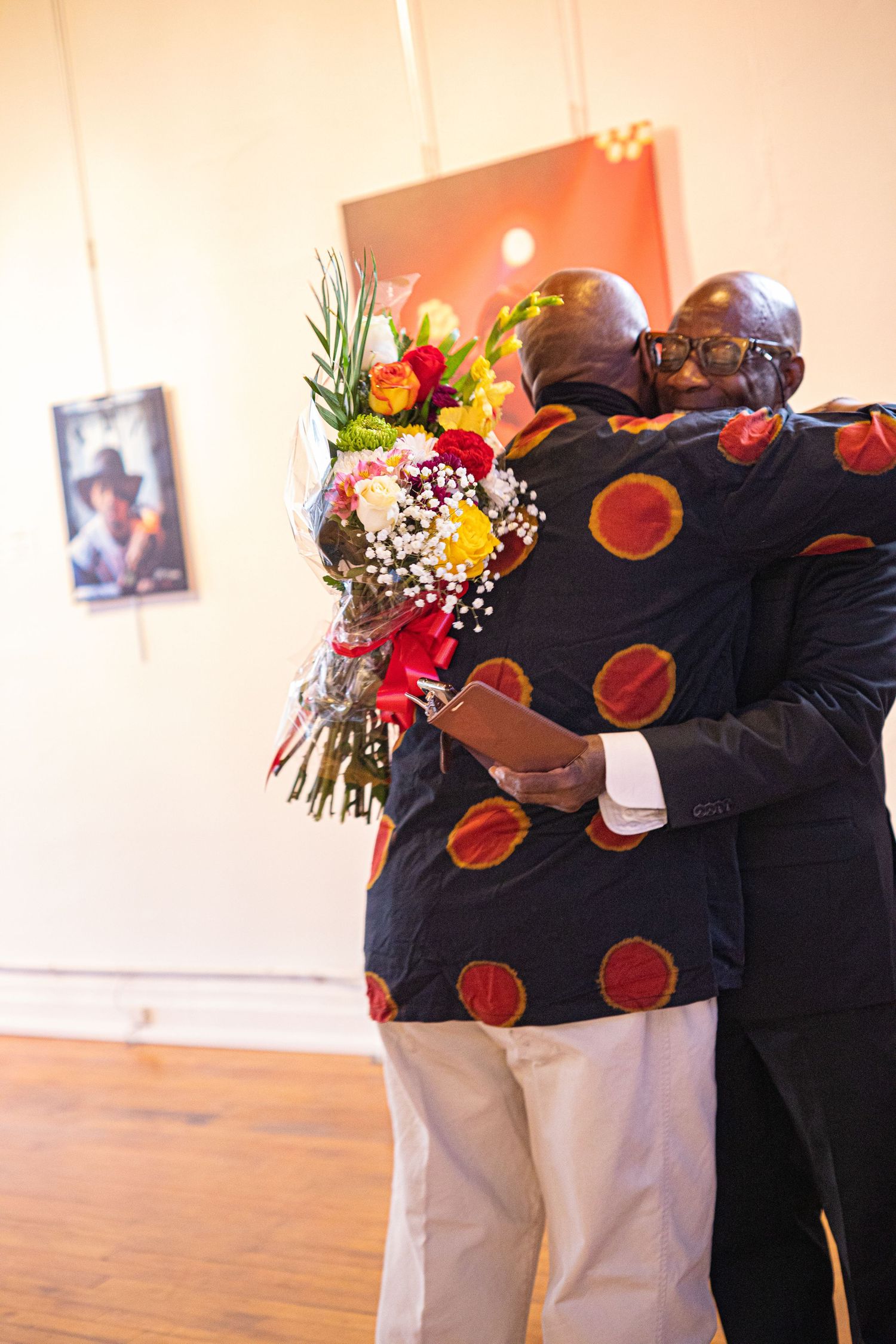 Two people embracing warmly in an art gallery, one holding a colorful bouquet of flowers, both wearing matching African print jackets.
