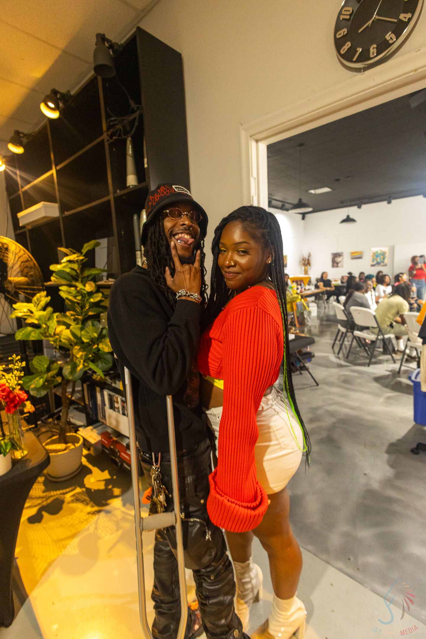 Couple poses together at indoor social gathering, man in black outfit and woman in red sweater.