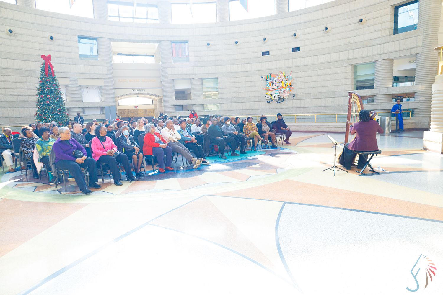 A harpist performs for a seated crowd in a rotunda decorated with a Christmas tree during a holiday concert.