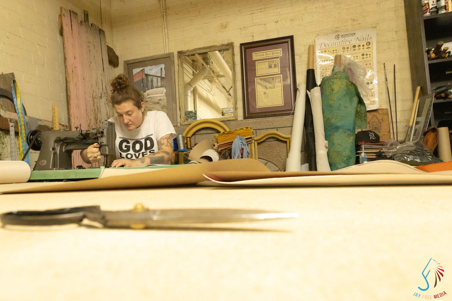 Woman working at a craft table in a workshop with tools and framed artwork visible in the background.
