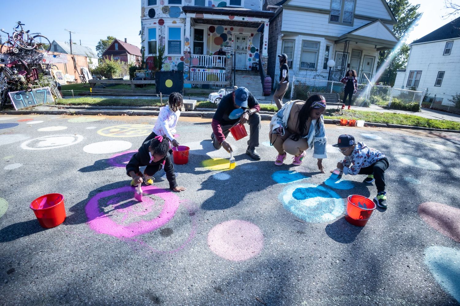 Children and adults paint colorful chalk circles on a street pavement with red buckets on a sunny day.