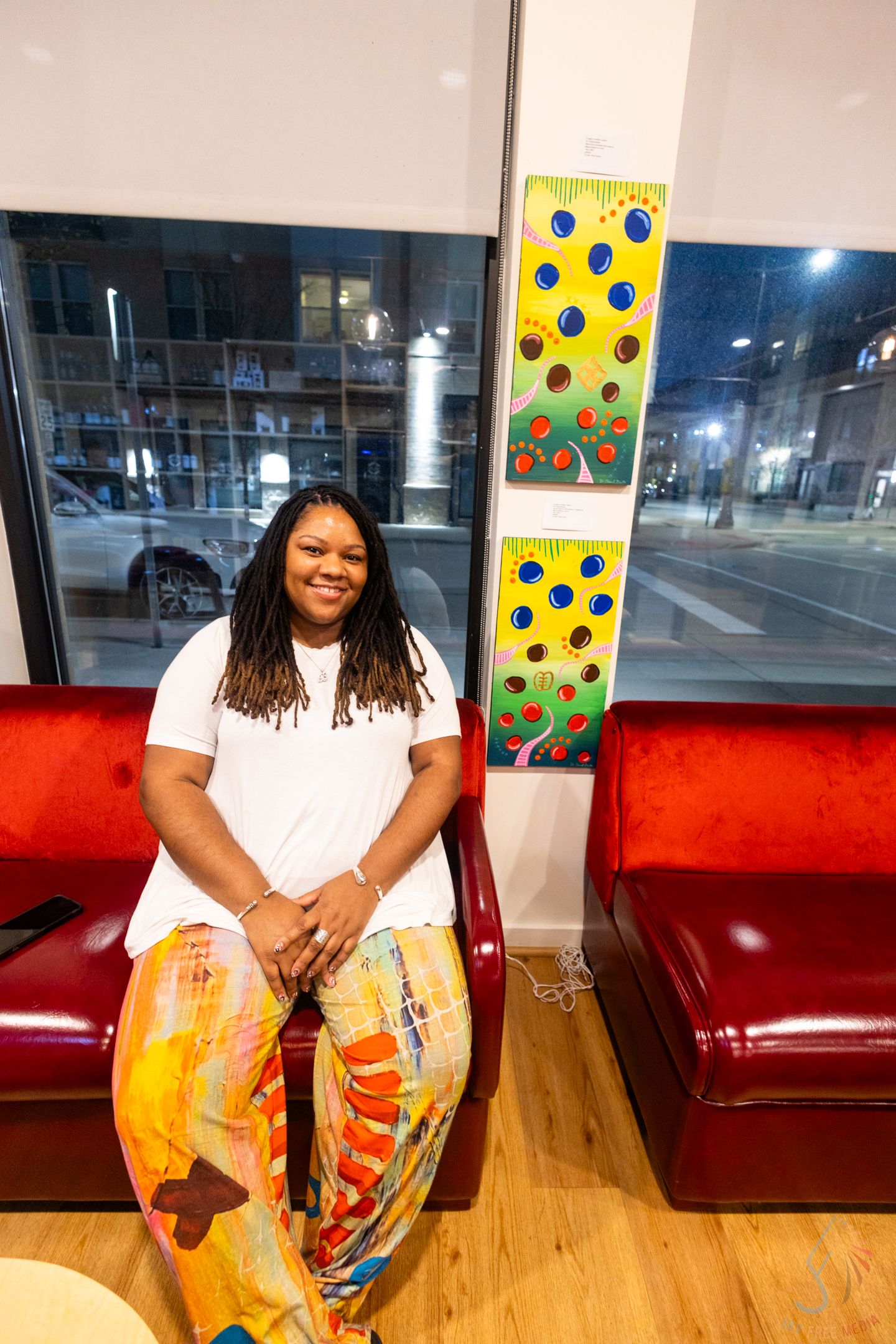 Woman with locs in white top and colorful pants sits smiling on red couch beside vibrant artwork in modern space.