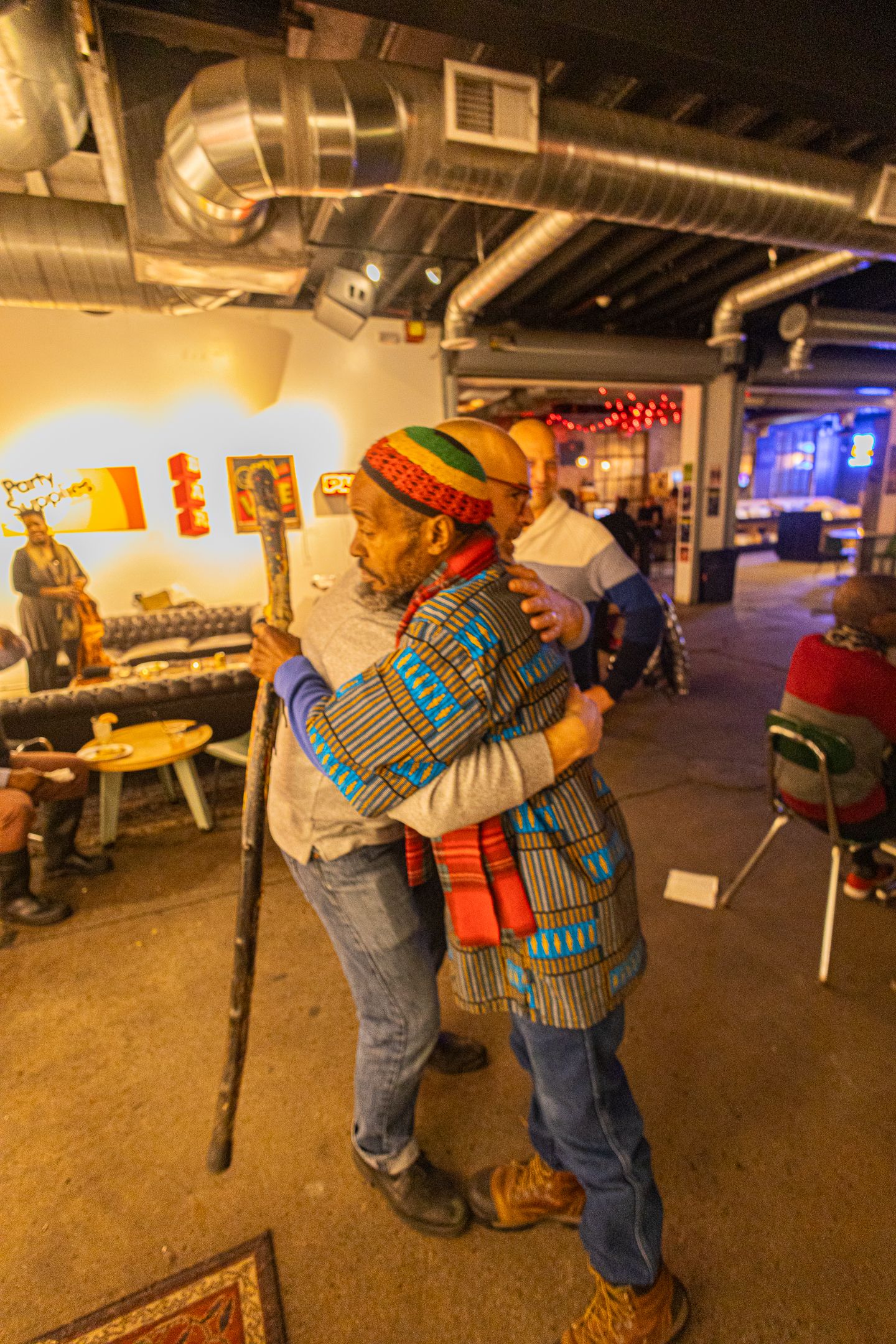 Two men in colorful African attire share a warm hug inside a vibrant indoor venue with exposed ductwork.