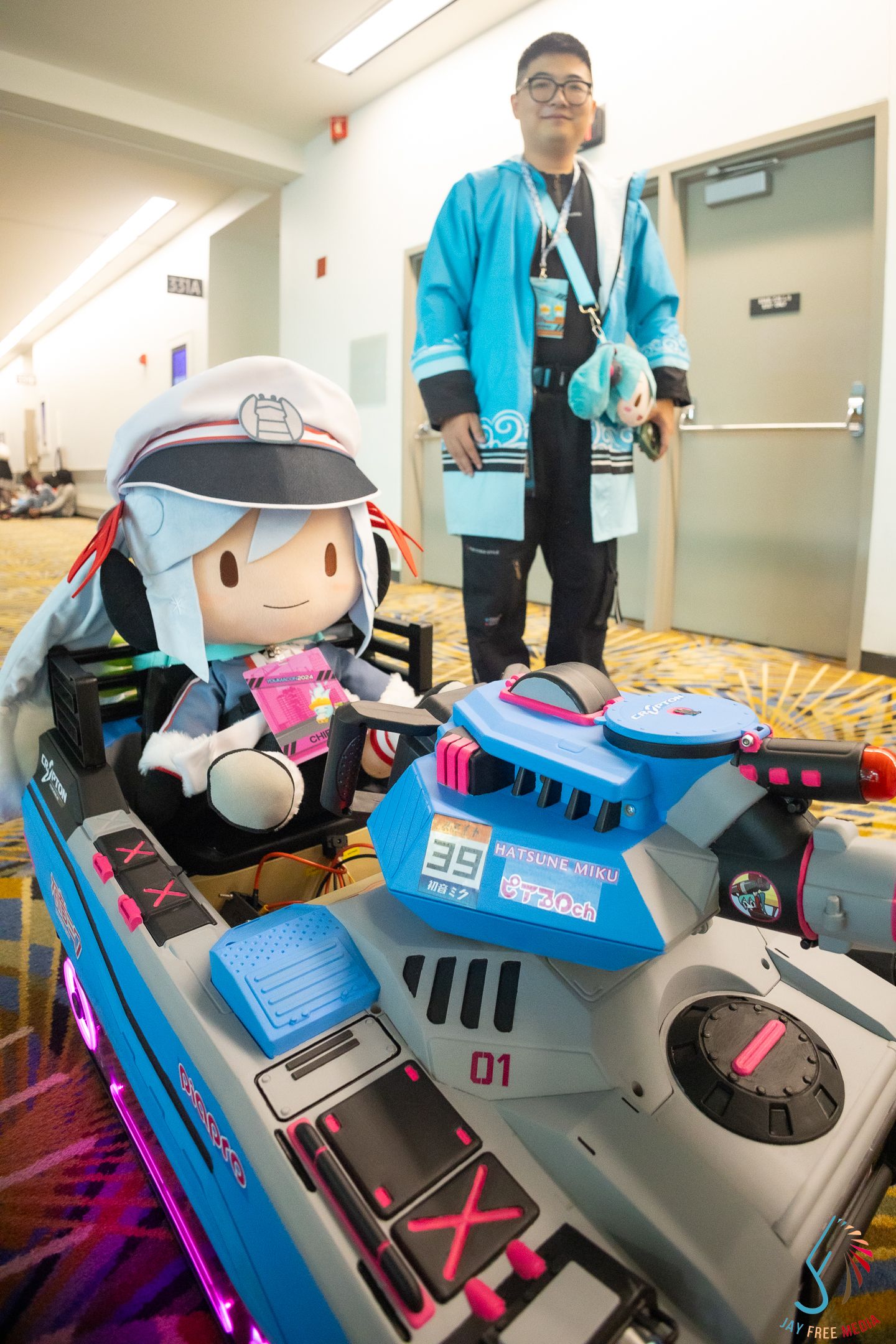 Close-up of D.Va mech cosplay prop with plush figure, cosplayer in teal outfit standing behind.