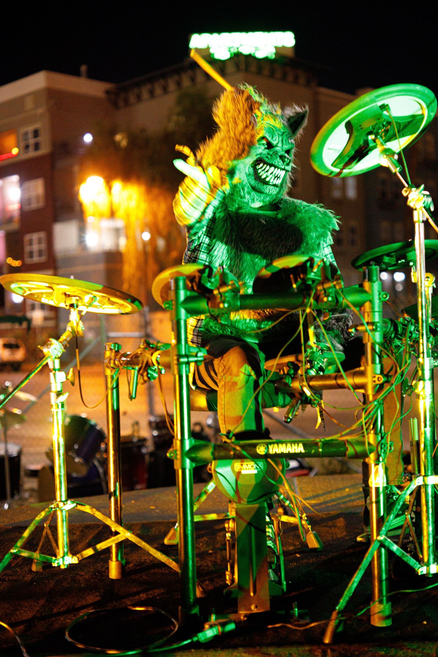 A performer in a green monster costume plays a Yamaha drum kit under dramatic green lighting at a nighttime outdoor event.