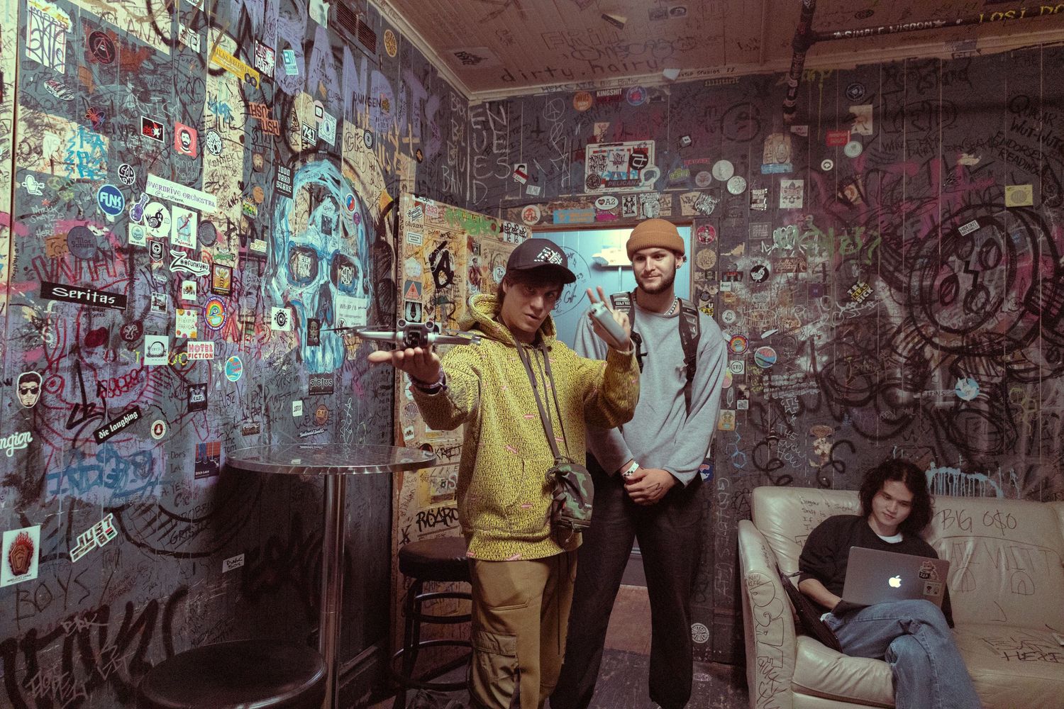 Three young people in a graffiti-covered room, two standing and one sitting on a couch with a laptop.