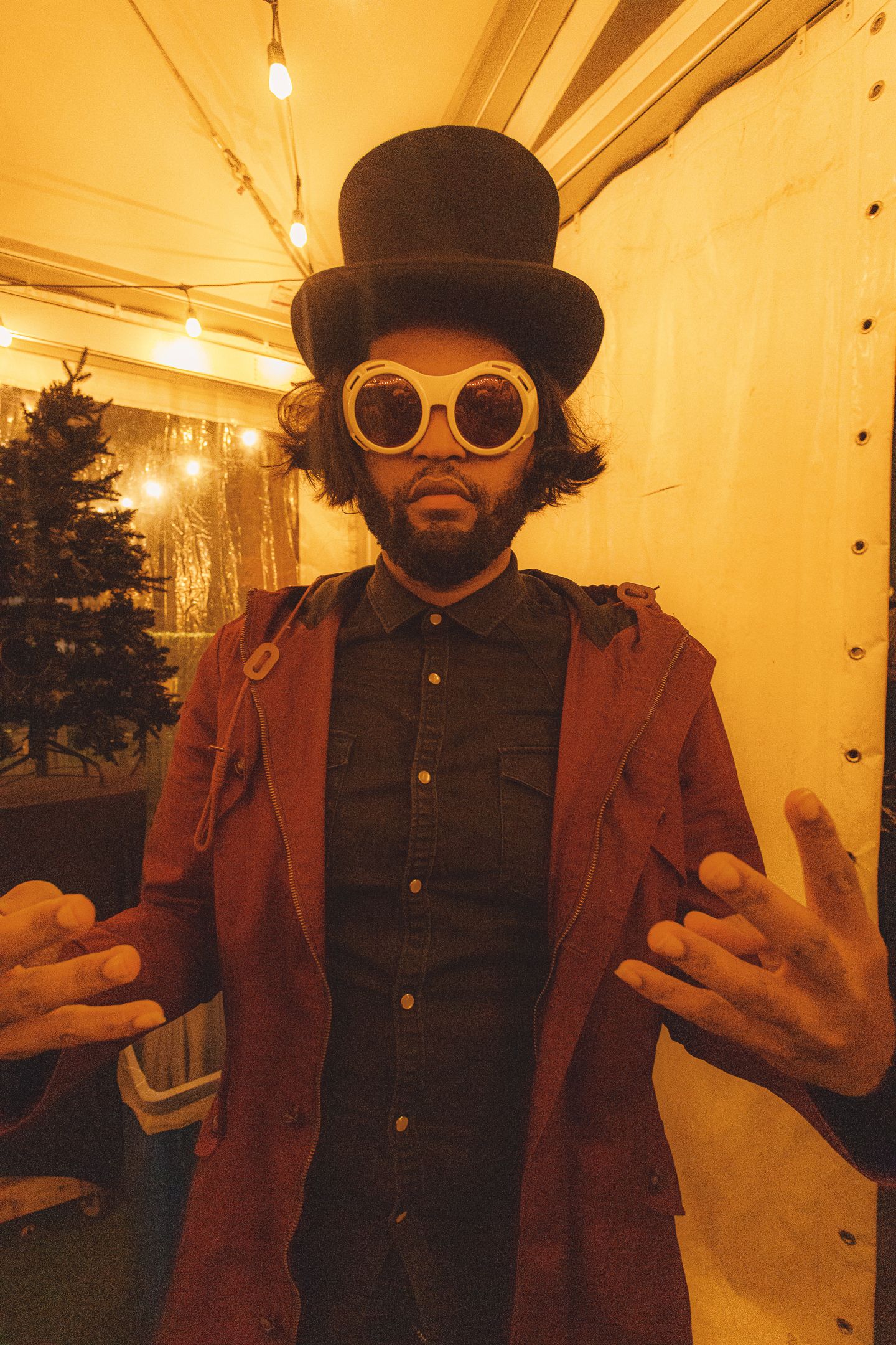 Man in black top hat, round white goggles, and red coat posing dramatically in warmly lit festive setting.