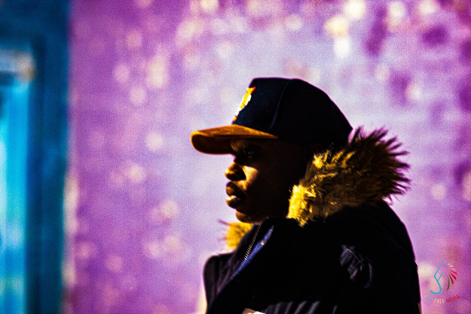 Person wearing a snapback cap and fur-trimmed jacket against a vibrant purple bokeh background.