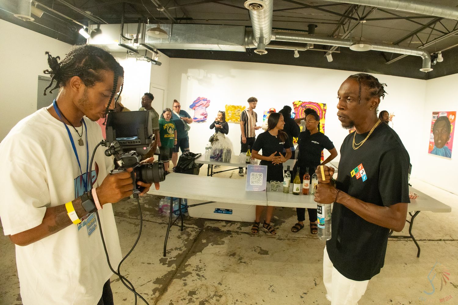 Videographer filming a man in white tee at an indoor art gallery event with people in background.