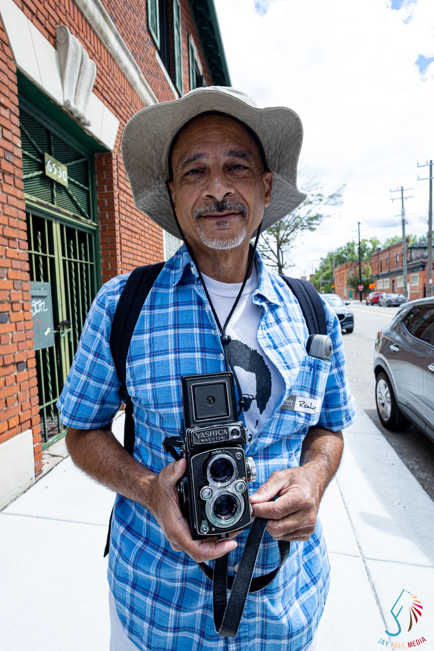 Man in blue plaid shirt and sun hat holds vintage Yashica twin-lens camera outdoors.