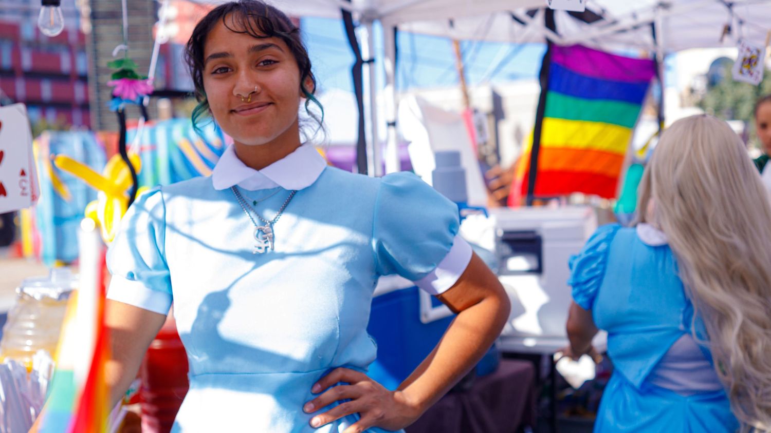 Young woman in light blue dress poses at outdoor festival with rainbow flag in background.