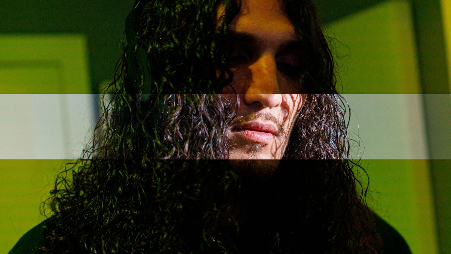 Person with long dark curly hair posing against a green wall in dramatic lighting.