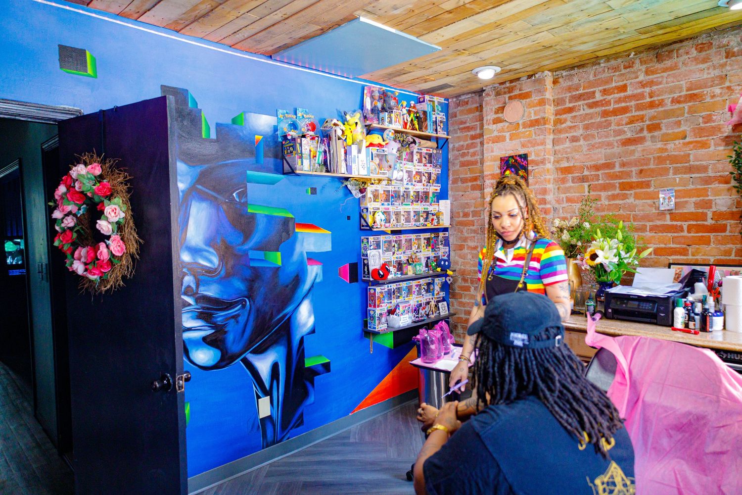 Nail technician works at vibrant salon with colorful mural, brick wall, and product display in background.