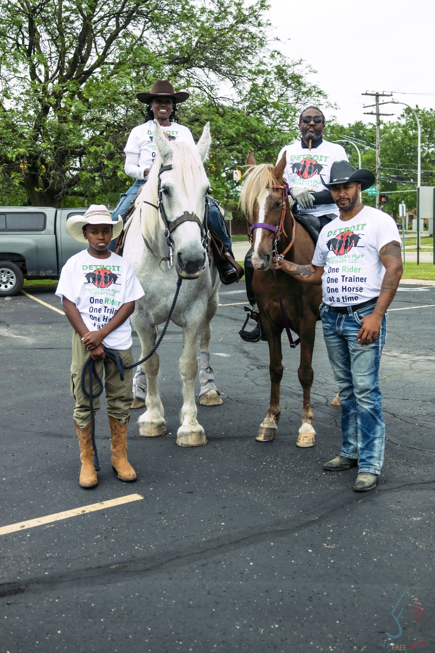 Group of cowboys in matching white shirts with two horses at an outdoor event in a parking lot.