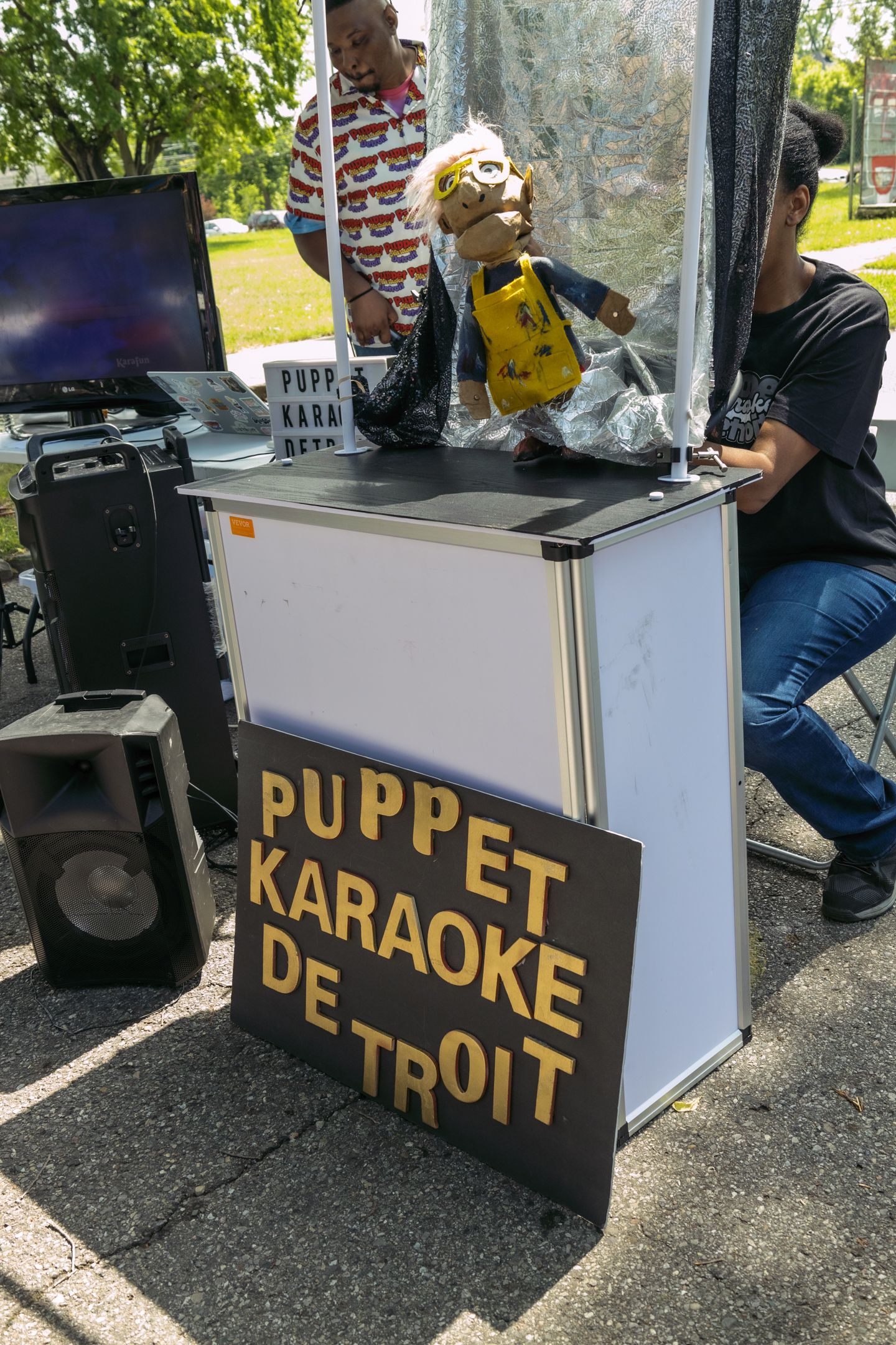 Puppet Karaoke Detroit booth with a puppet display, speaker, and sign at an outdoor community event.