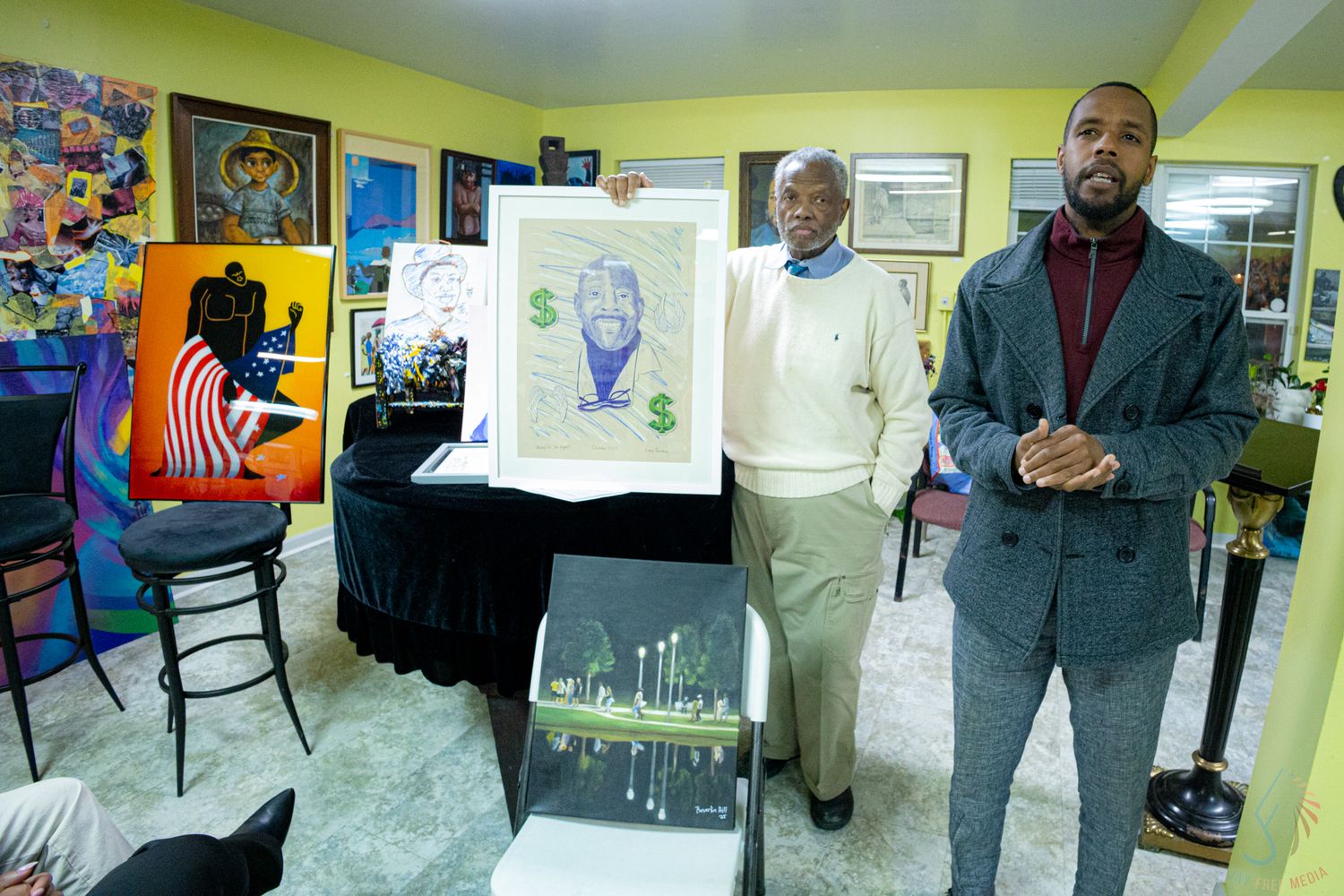 Two men display a green dollar sign artwork print at a colorful indoor art gallery.
