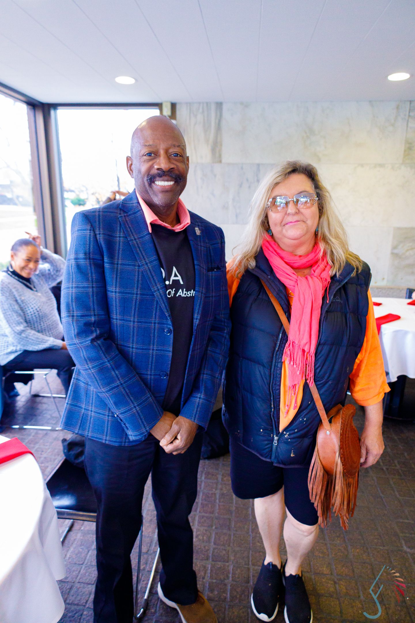 Two people smiling indoors, man in blue plaid blazer and woman in orange scarf posing together.