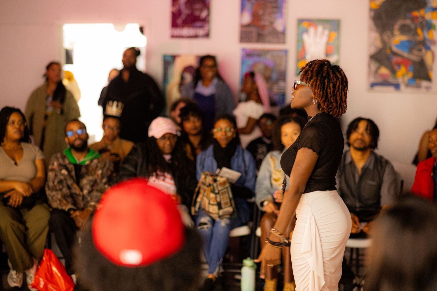 A speaker addresses a diverse seated crowd in an art-filled room during a community gathering or cultural event.