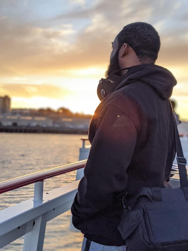 Person in hoodie watching sunset from boat deck over urban waterfront.