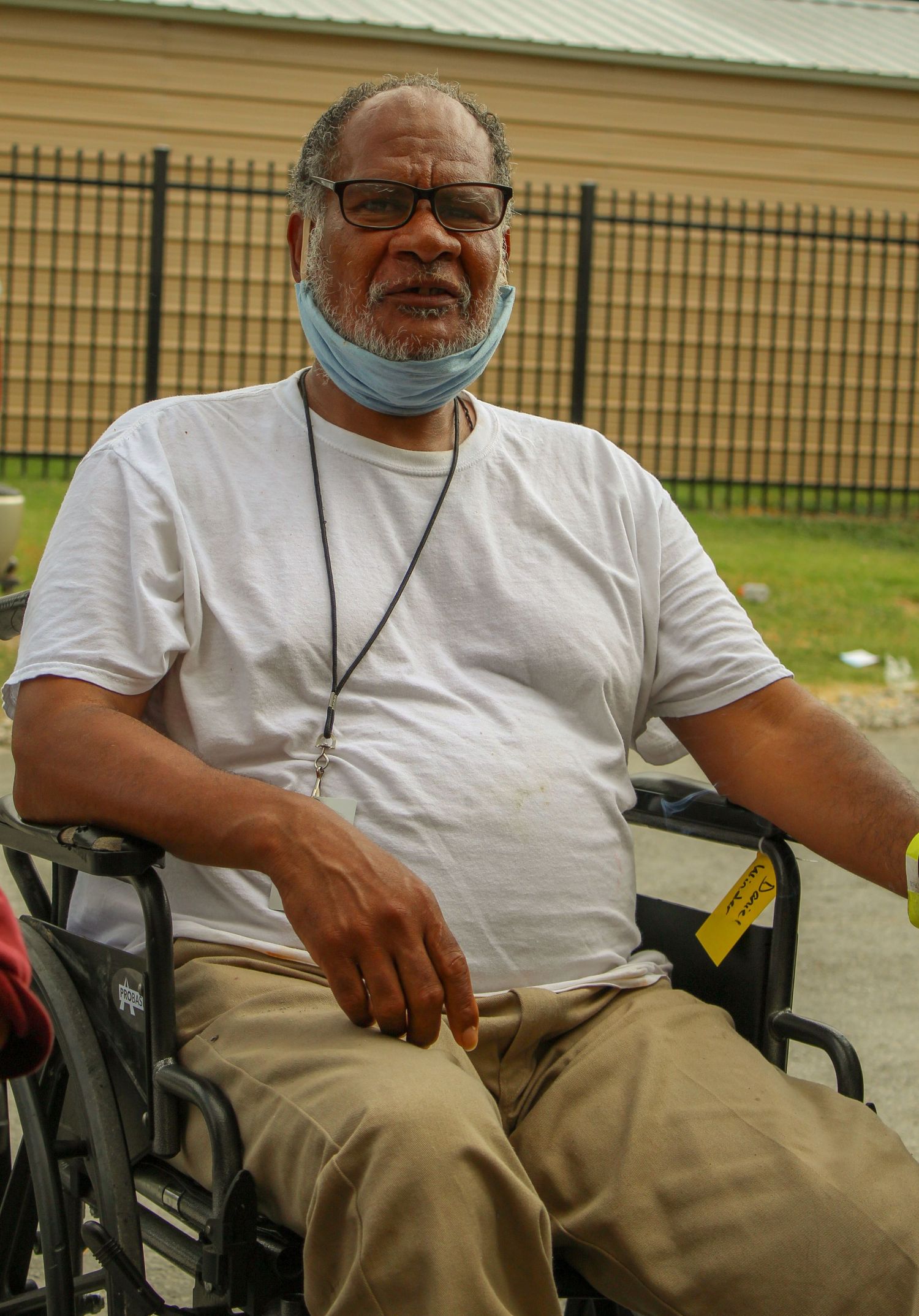 Person in wheelchair wearing white t-shirt and face mask sitting outdoors.