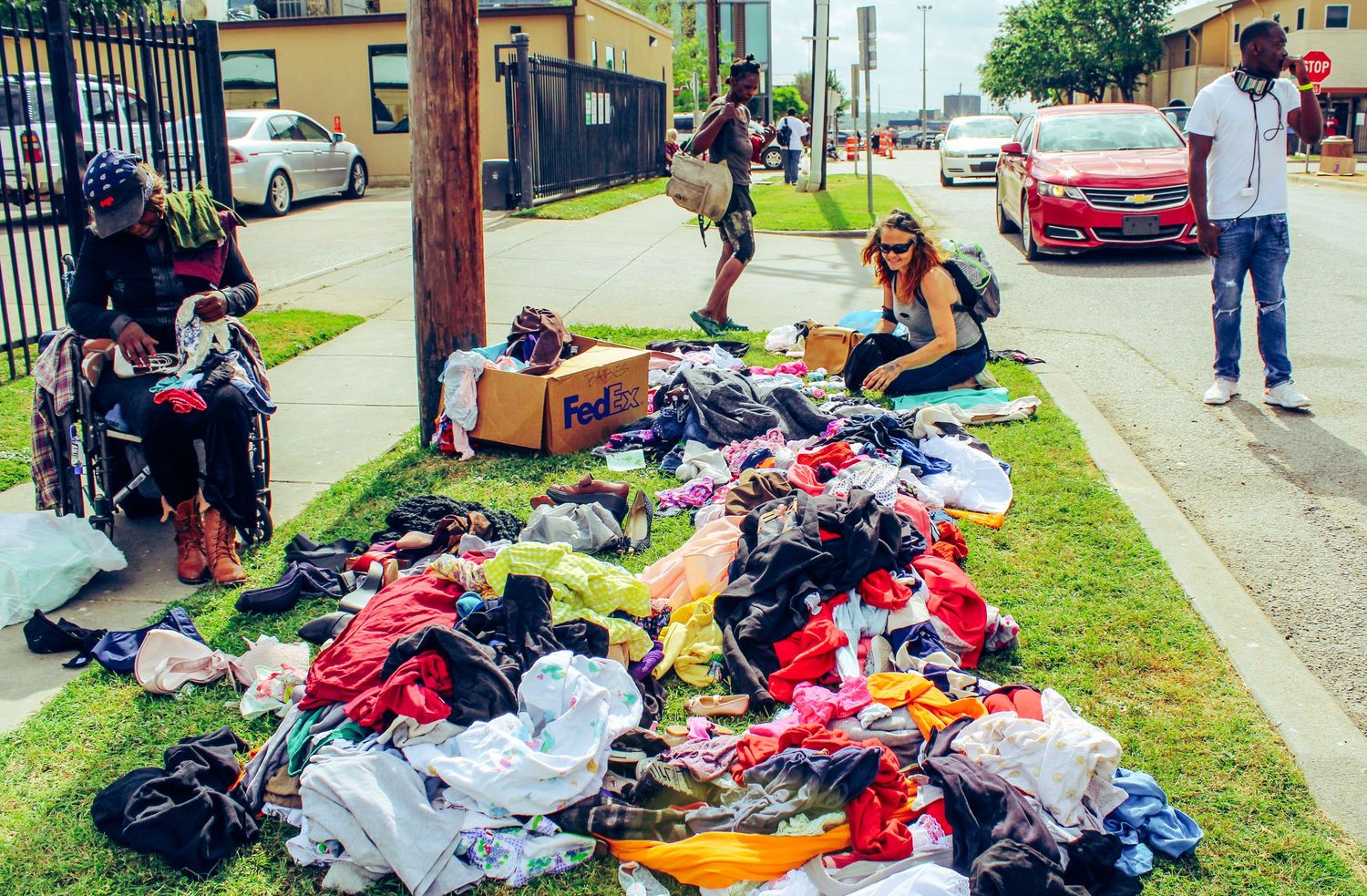 Piles of clothing and items spread out on grass beside a sidewalk.