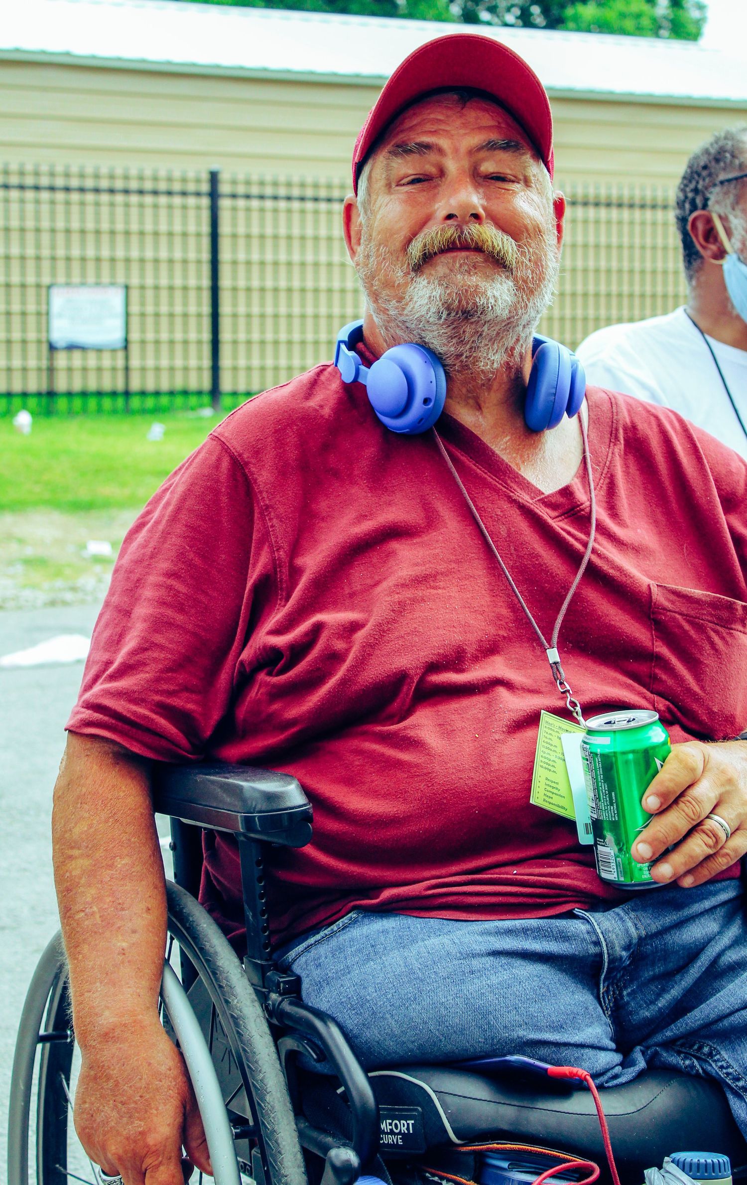 Person in wheelchair wearing red cap and shirt with blue headphones holding beverage can outdoors.