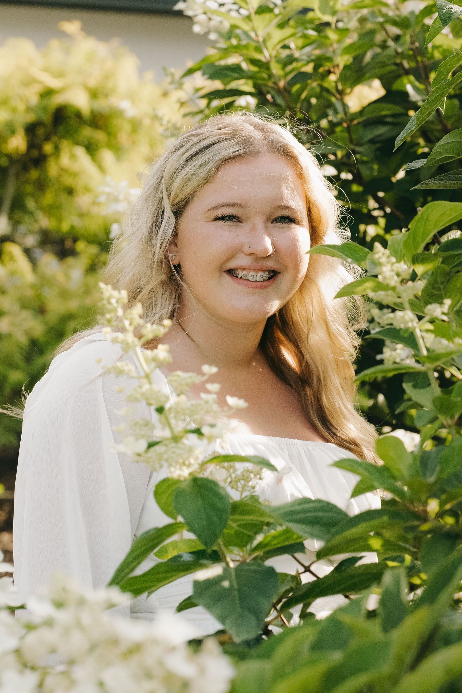 Smiling blonde teen with braces in white top surrounded by blooming white hydrangea flowers in a sunny garden.
