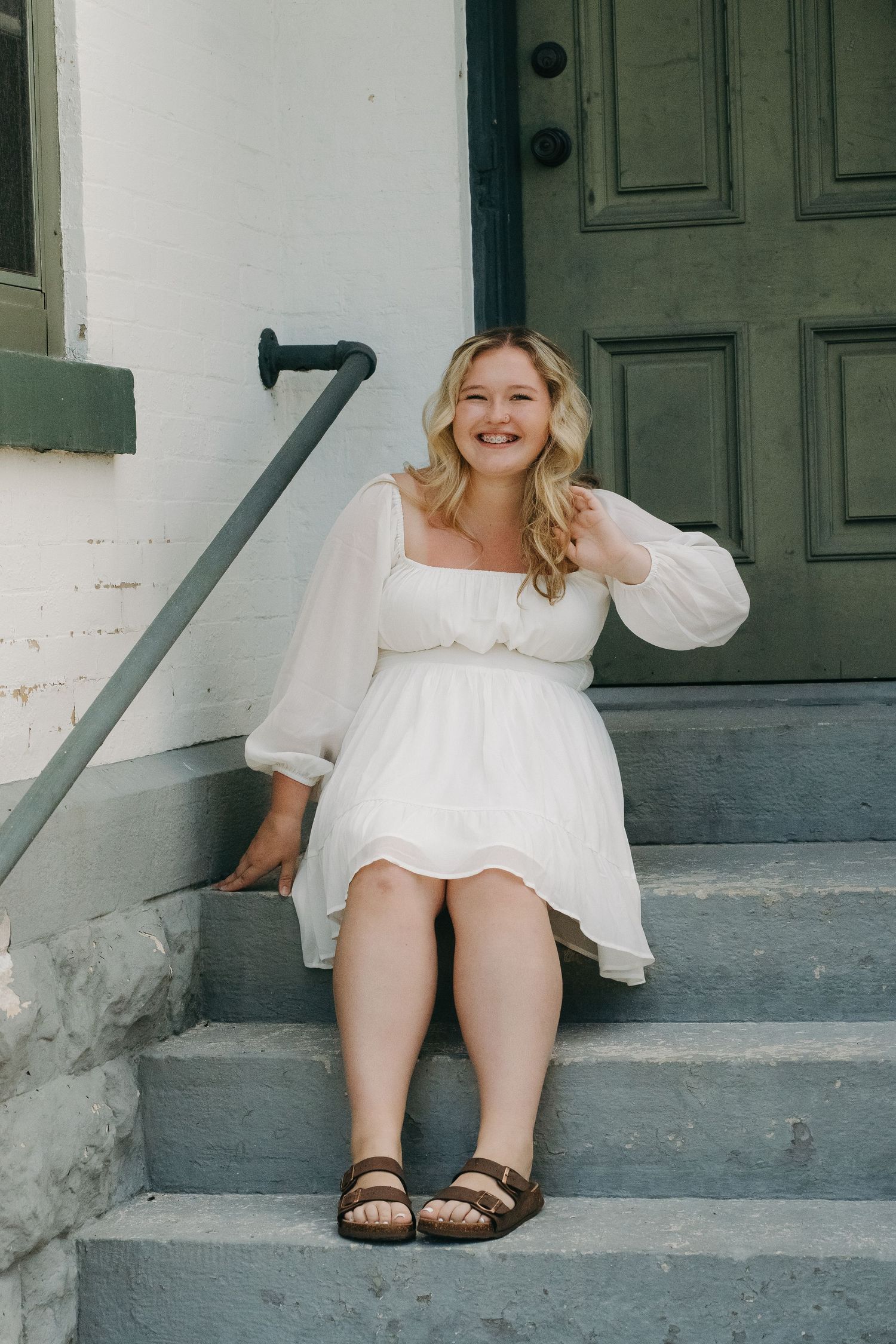 Blonde woman in white dress sitting on gray steps by green door.